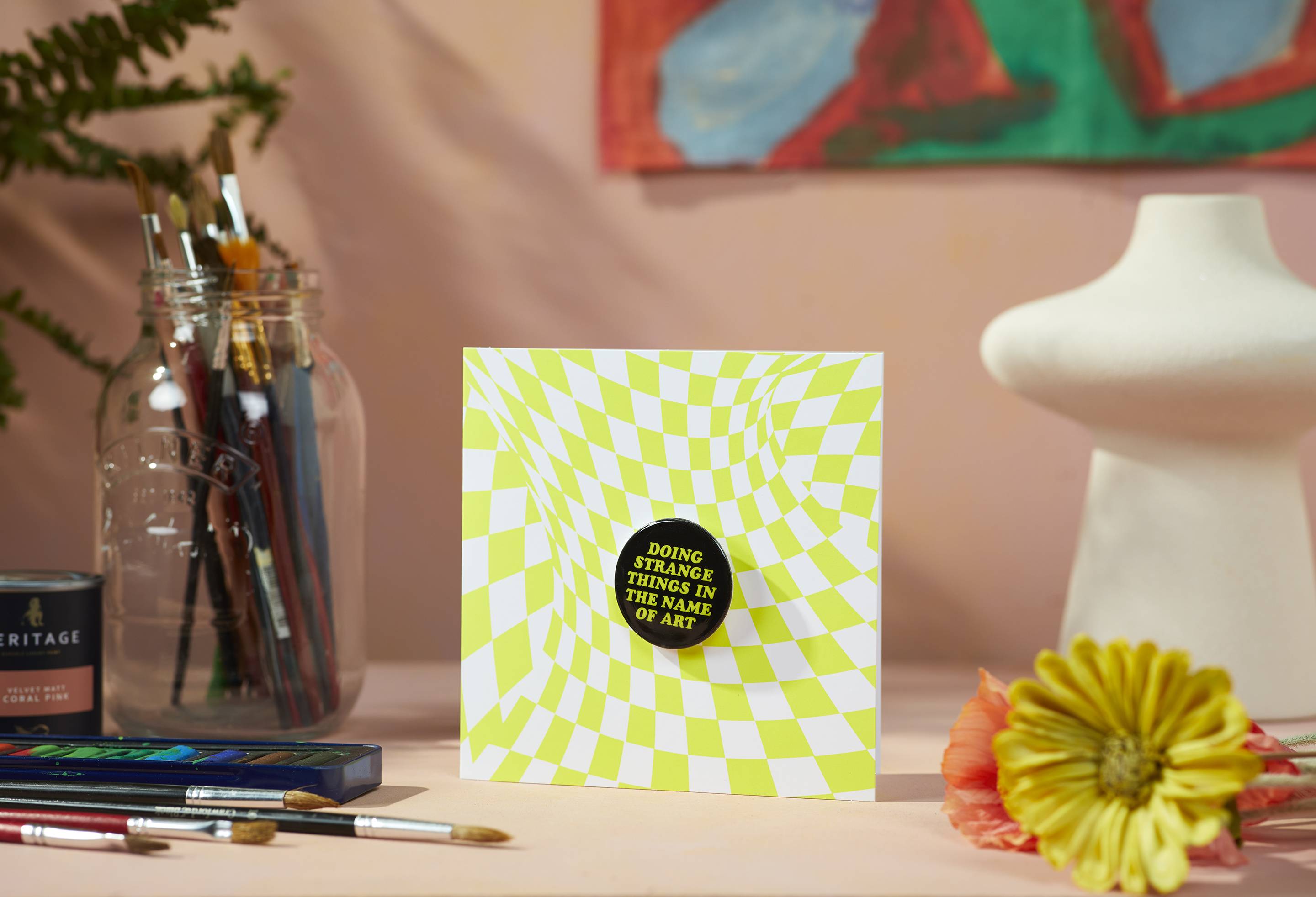 The funky-patterned greetings card is sitting on a pastel table surrounded by a sculpture, flowers and a multitude of paintbrushes.