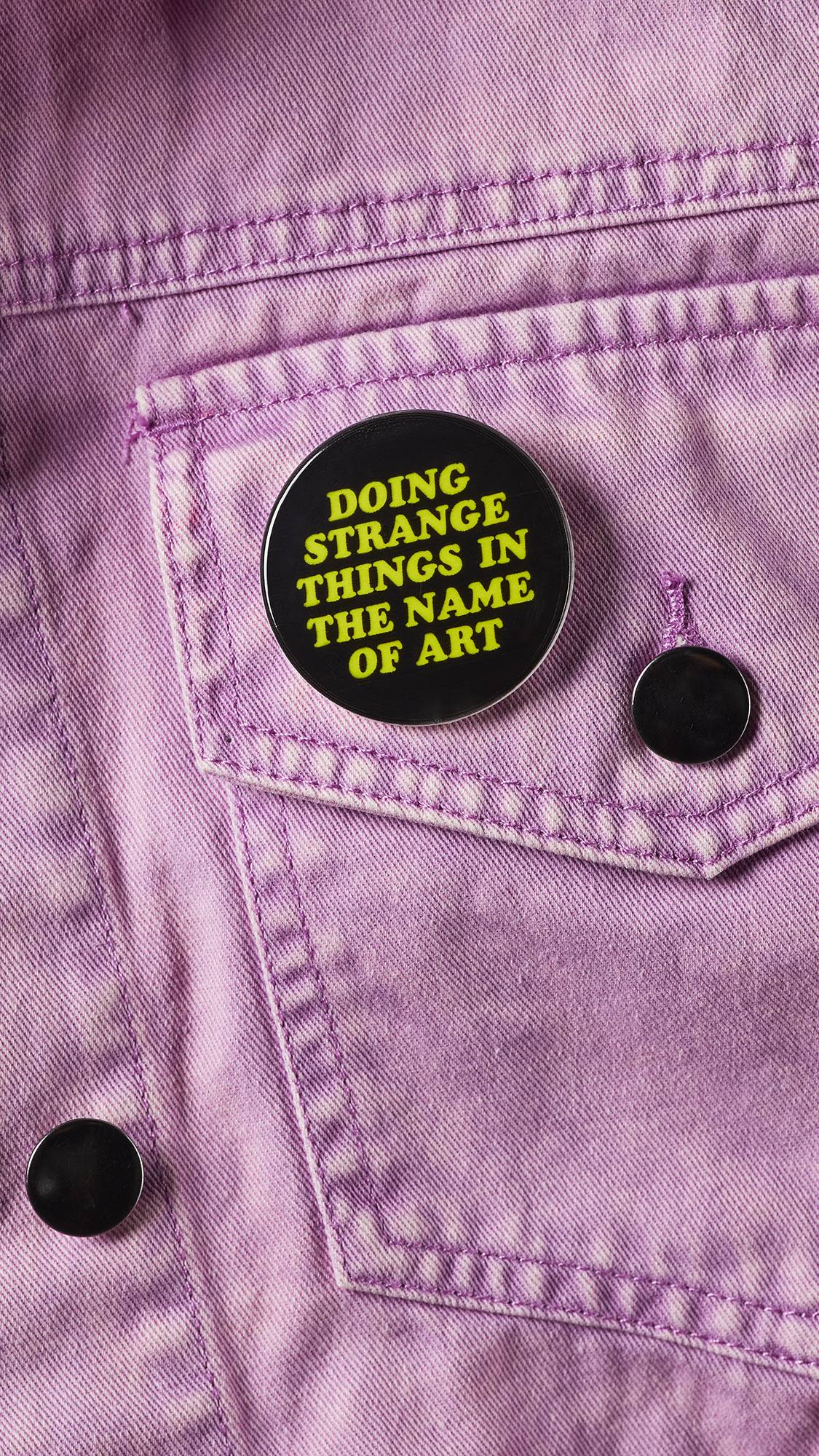 A close-up of the black badge with the green text "Doing Strange Things In The Name Of Art" pinned to a baby pink denim jacket pocket. 