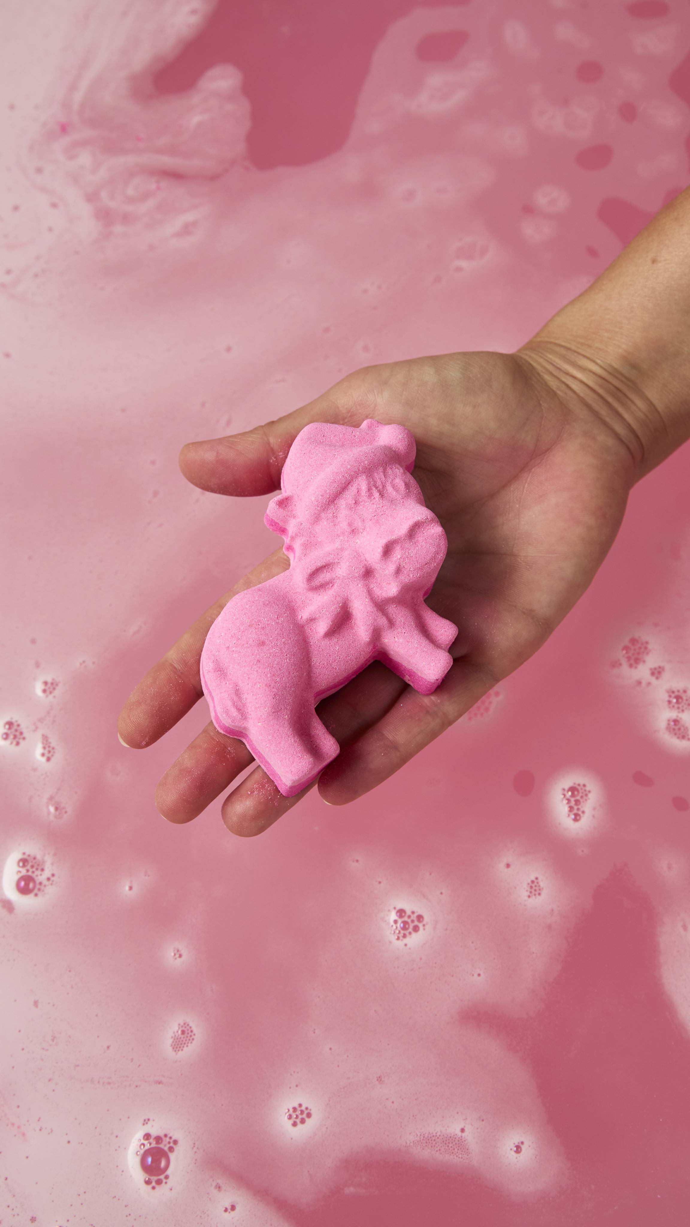 A hand holds a vibrant pink donkey-shaped bath bomb wearing a hat over swirling, pastel pink bathwater topped with soft white bubbles.
