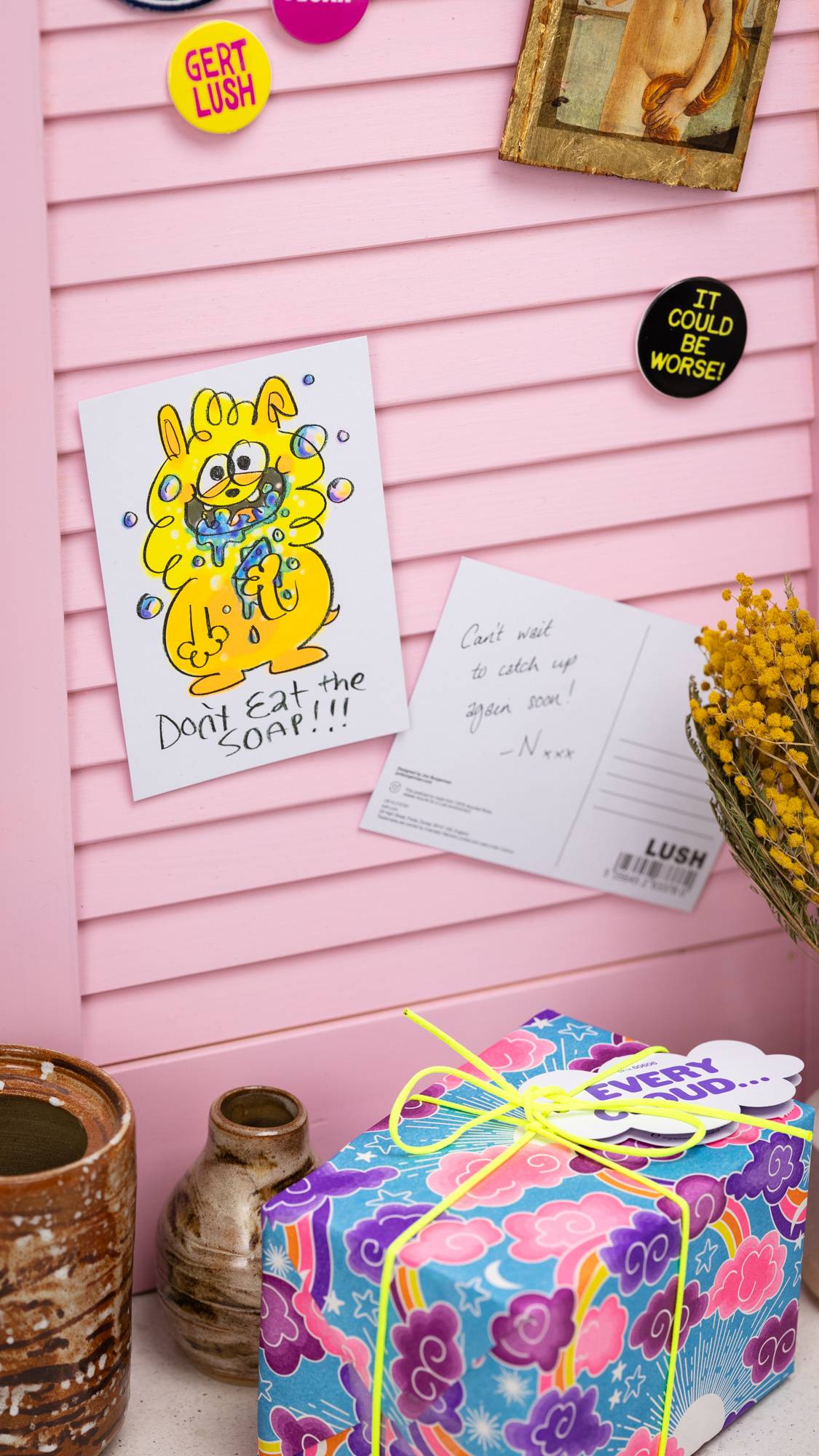 Image shows the Don't Eat The Soap postcard on a pastel pink noticeboard surrounded by badges and a gift.