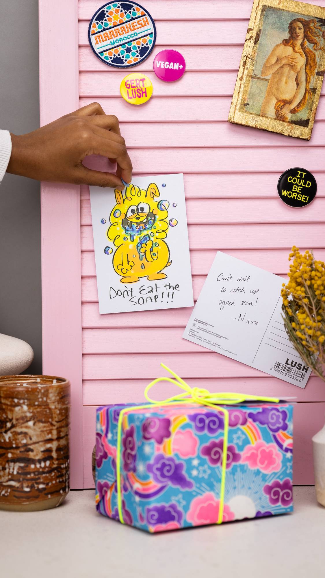 Model is pinning the Don't Eat The Soap postcard on a pastel pink noticeboard surrounded by badges and a gift.