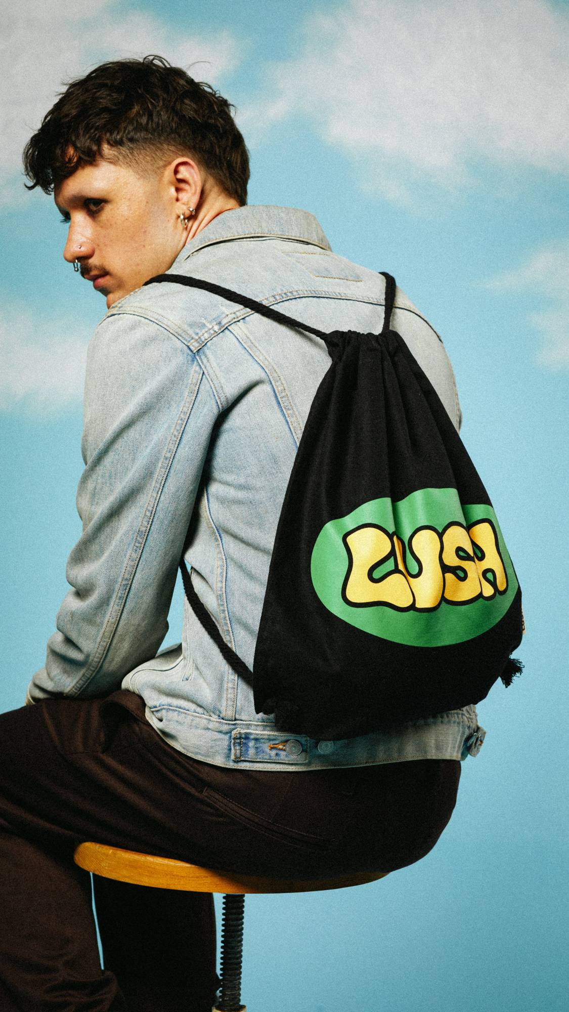 Model in on a sky-like background wearing a denim jacket, sitting on a stool wearing the Retro Bubble drawstring bag.
