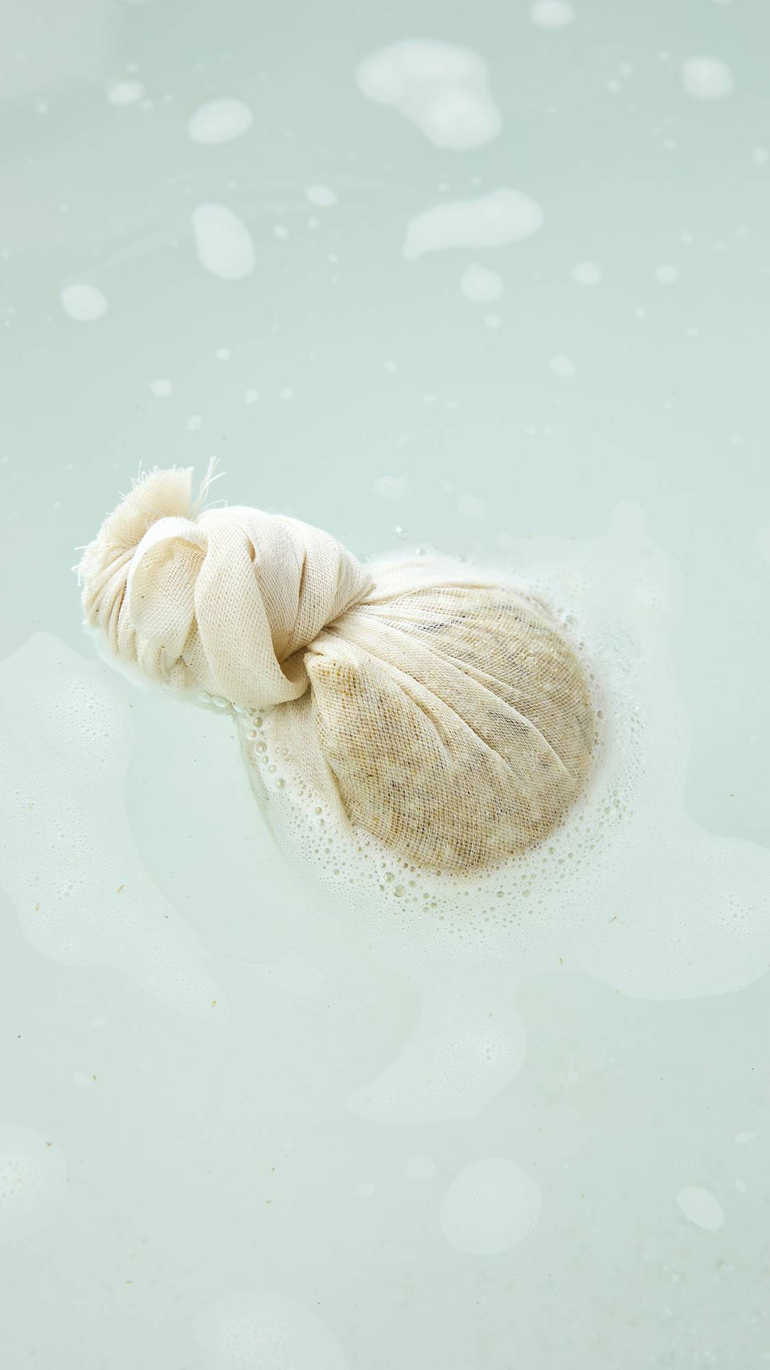 An aerial view of a Lush "Dream Cream Soak" bath bomb wrapped in a cream muslin cloth pouch floating in warm bathwater, surrounded by soft white foam and gentle bubbles.