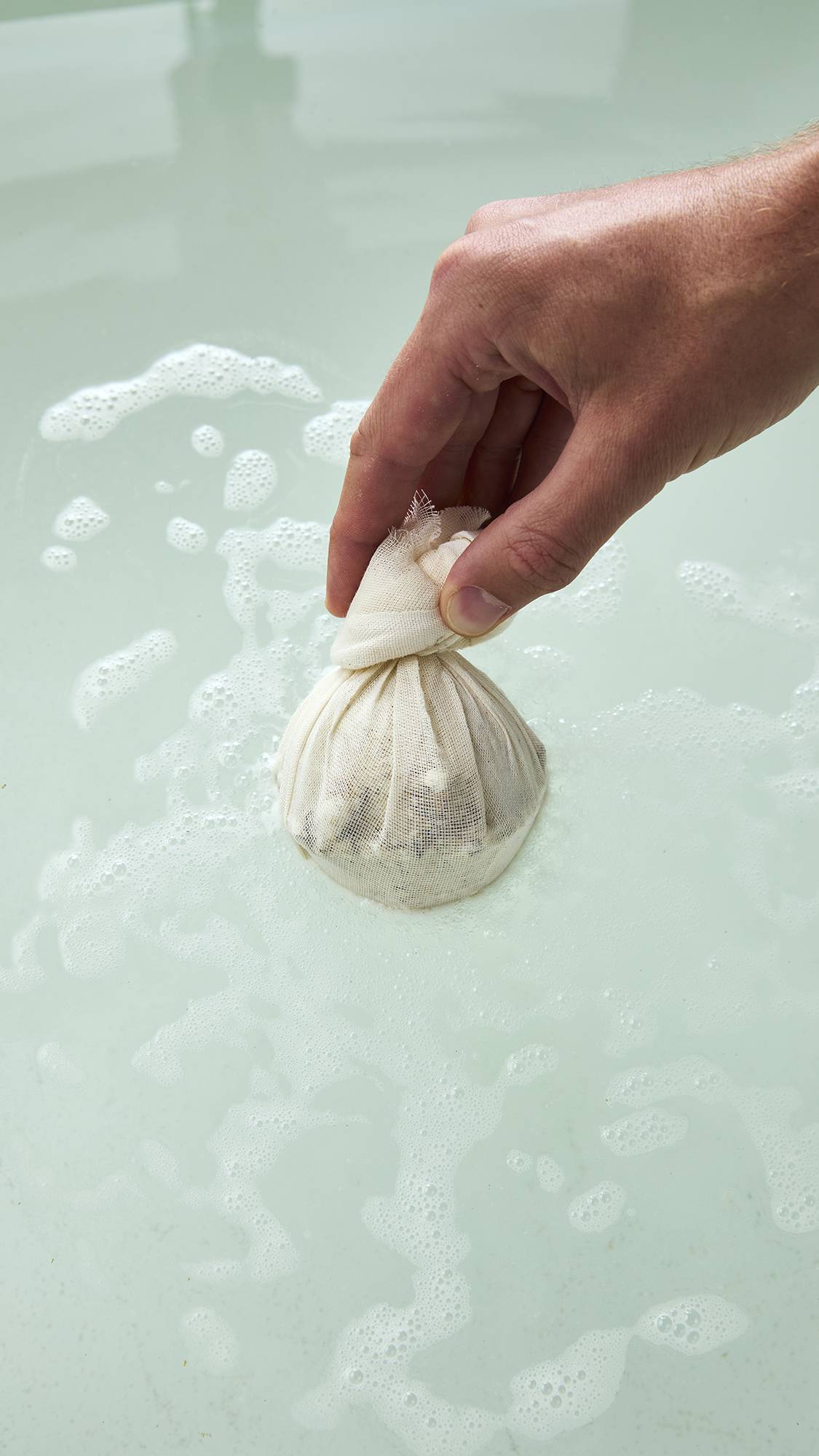 The image shows a hand lowering a LUSH "Dream Cream Soak" bath bomb wrapped in a cream muslin cloth pouch into warm, bubbly bathwater.