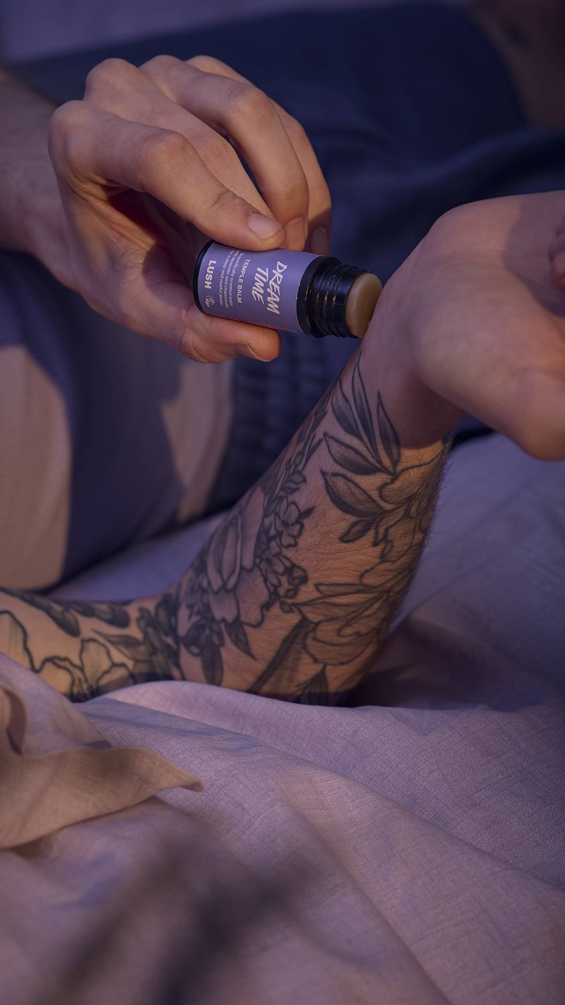 The image shows a close-up of the model's forearms and hands as they are lying in bed in lavender-purple sheets. They are gently swiping the temple balm across their wrists.