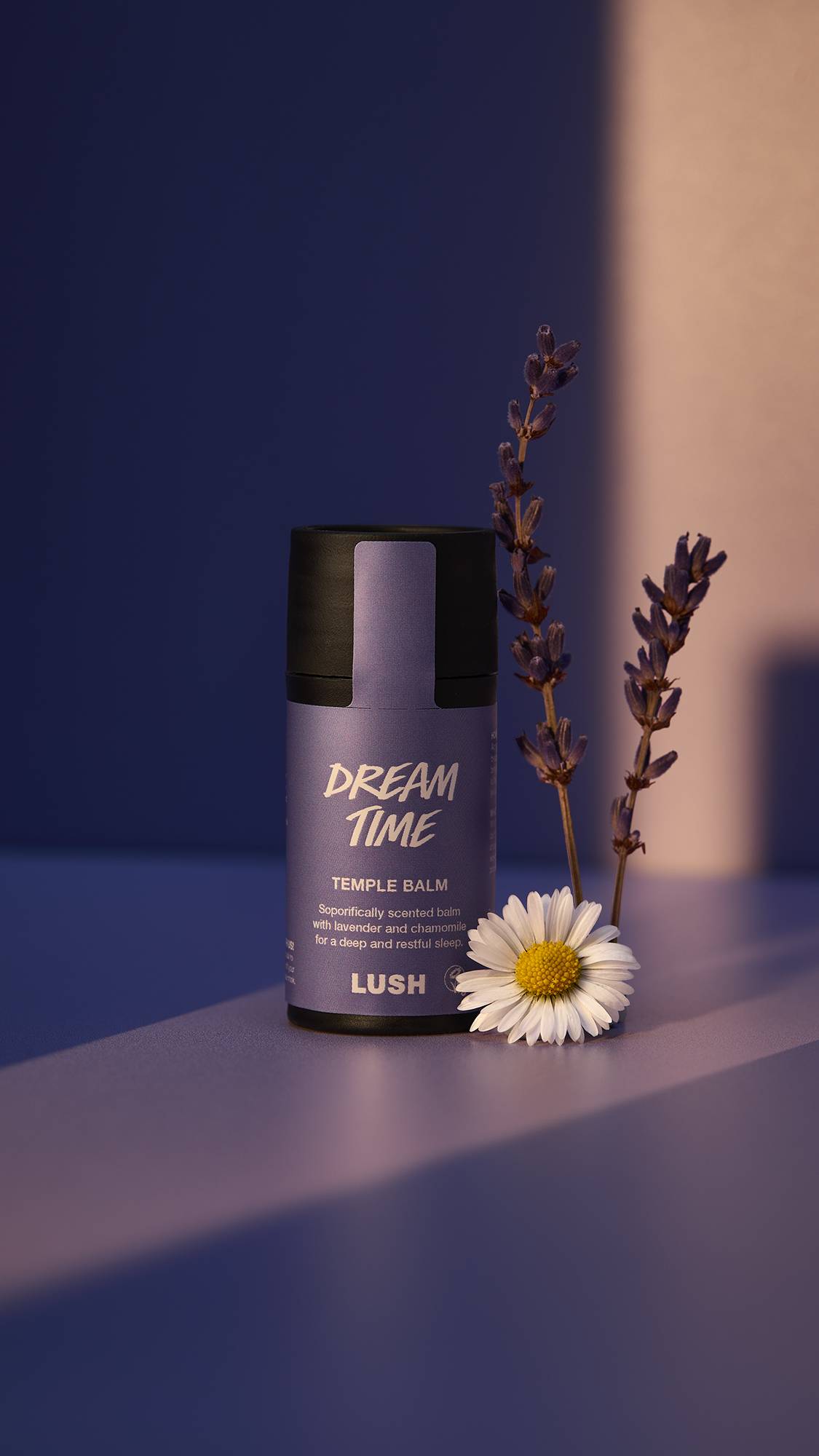 A close-up of the Dream Time temple balm surrounded by purple lighting. There is a single sprig of lavender and a single chamomile flower beside the product. 