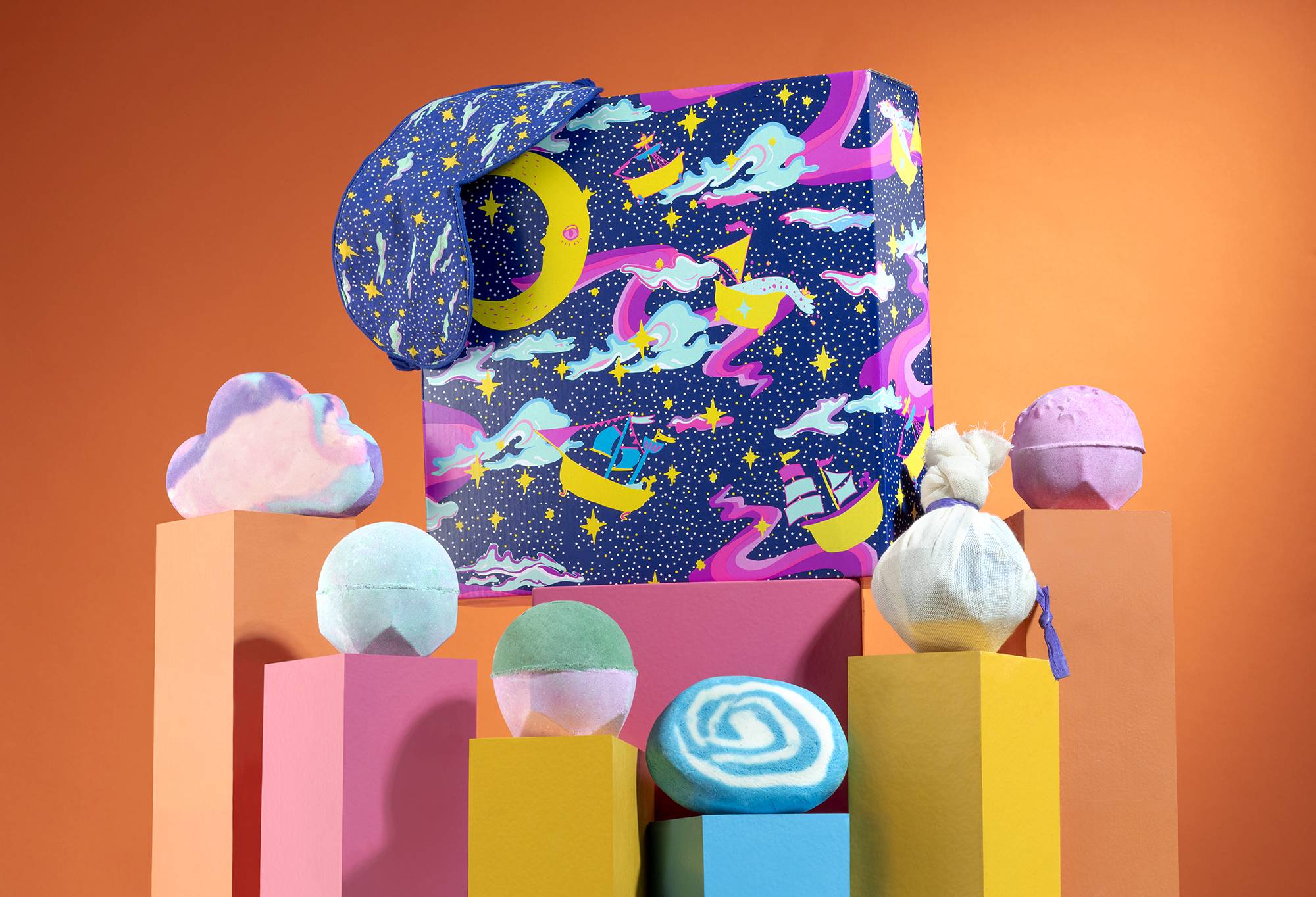 Dreamland gift, in front of an orange background, surrounded by its products on pink, orange, yellow and blue plinths.