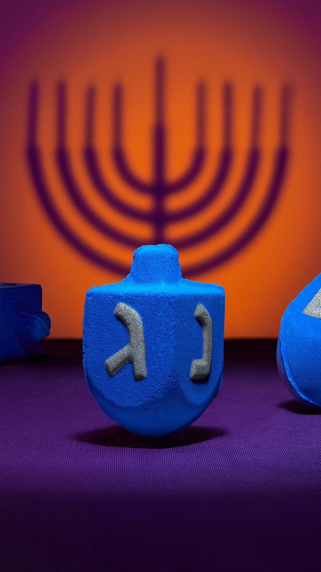 The image shows the Dreidel, Dreidel, Dreidel bath bomb propped up focusing on the details of the embossed characters on the side. The background is a deep orange and purple colour with the shadow of a Menorah. 