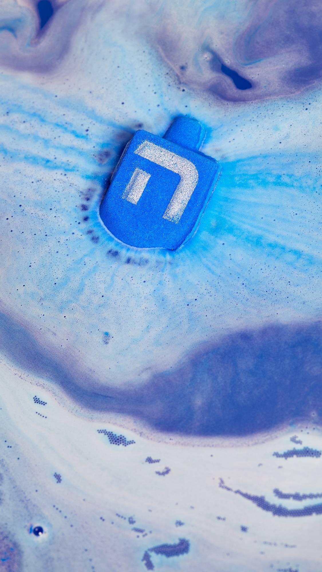 An aerial view of the Dreidel, Dreidel, Dreidel bath bomb as it floats on the surface of the bath water. It is giving off rippling ribbons of velvety blue and purple foam. 