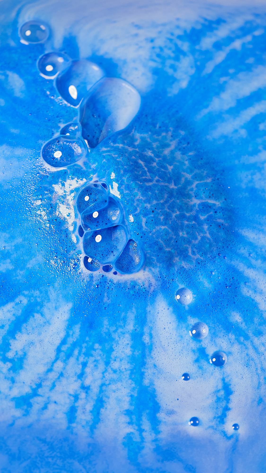 The image shows an aerial view of the final moments of the Dreidel, Dreidel, Dreidel bath bomb as it has almost fully dissolved. There is a blanket of thick, velvety, blue foam across the surface with a trail of large bubbles. 