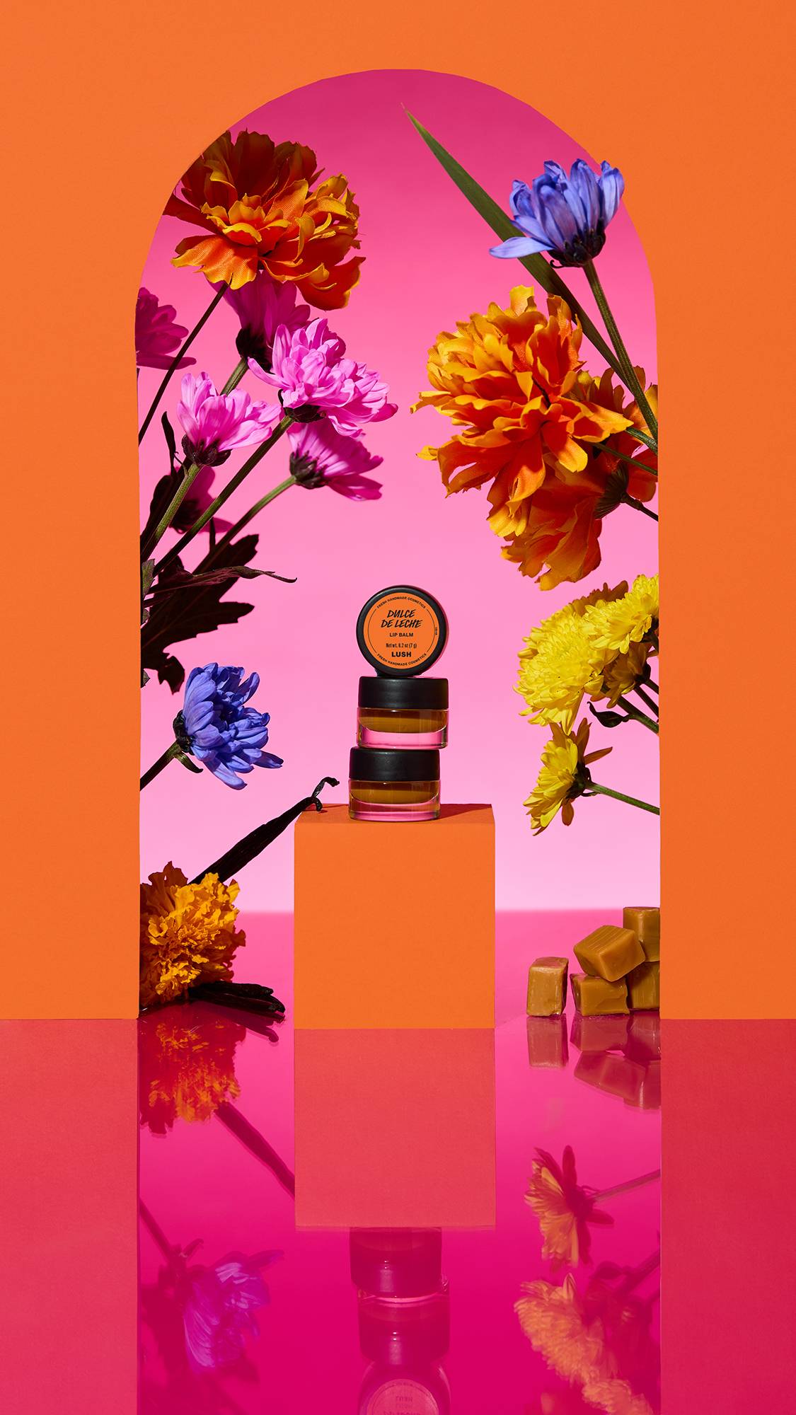 The image shows three stacked small glass jars of the Dulce De Leche lip balm on a plinth. A colourful arch frames the scene, with sliced citrus, flowers, and a cinnamon stick surrounding the product on a reflective pink surface.