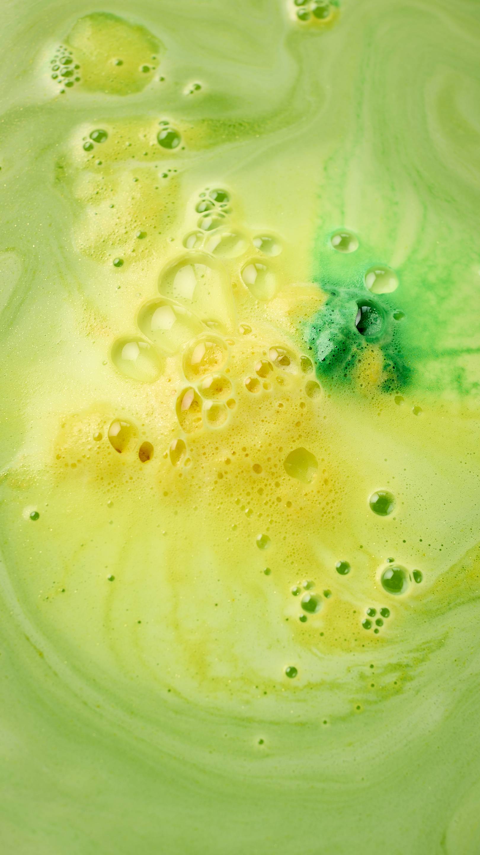 A gold-shimmering Easter Bunny bath bomb, shaped like a rabbit in a green egg, floats in vibrant neon green bathwater. It releases swirling trails of bright yellow foam across the surface. All glitter used is plastic-free.