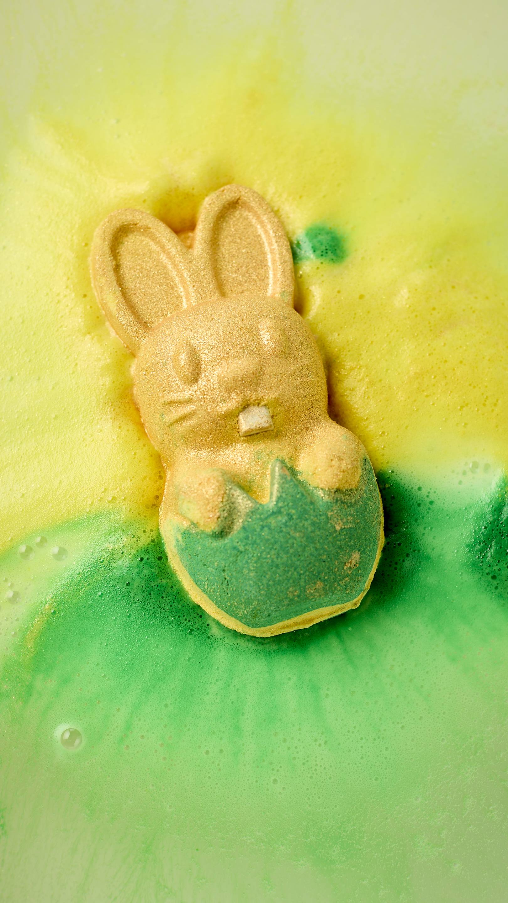 The Easter Bunny bath bomb, a golden rabbit popping out of a green grass-like base, dissolves in water. It releases vibrant trails of neon yellow and emerald green foam. Shimmering plastic-free glitter sparkles across the surface.