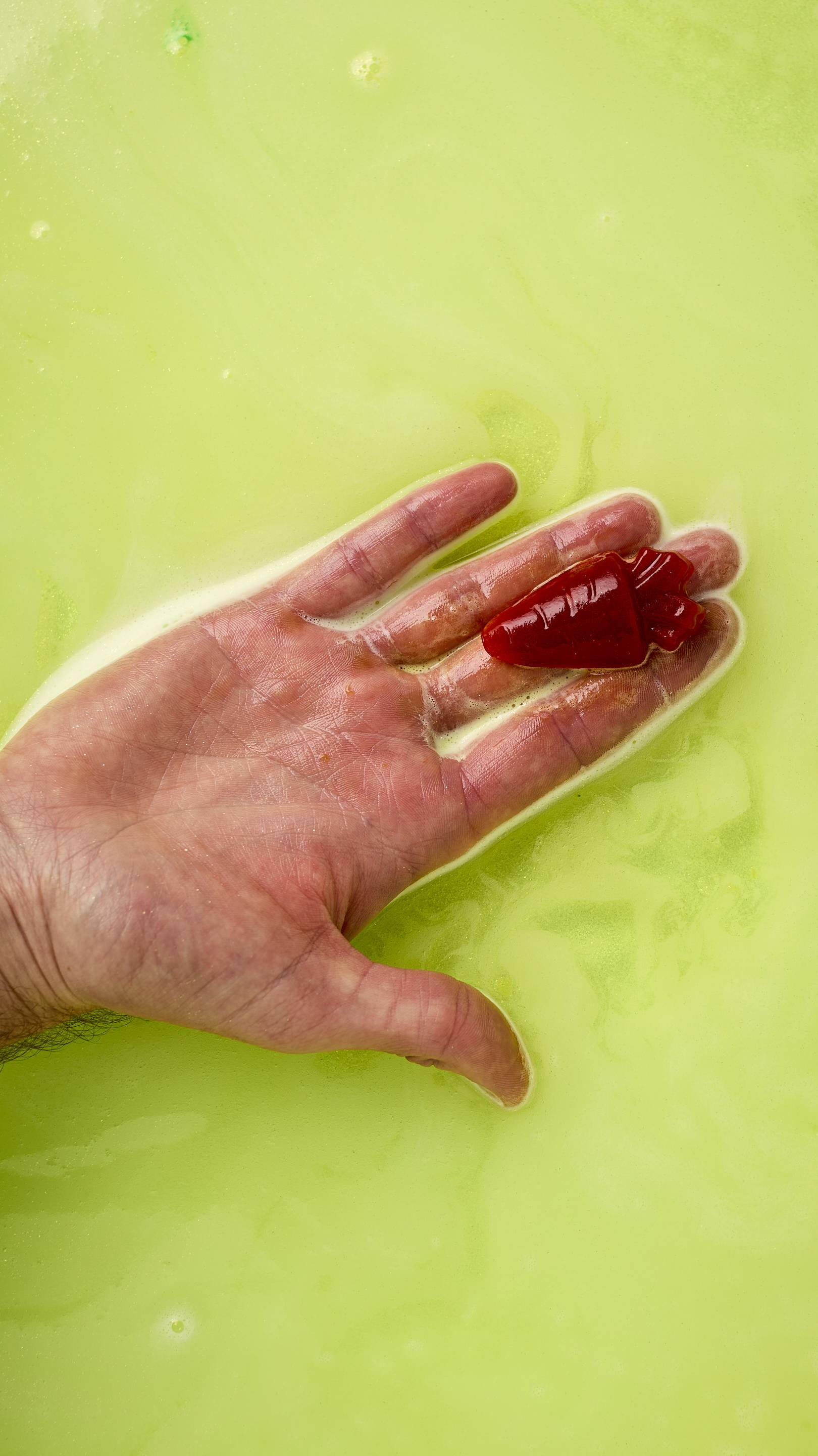A gold-shimmering Easter Bunny bath bomb, shaped like a rabbit emerging from a green egg, floats in vibrant neon green bathwater. It releases swirling trails of bright yellow foam across the surface. All glitter used is plastic-free.