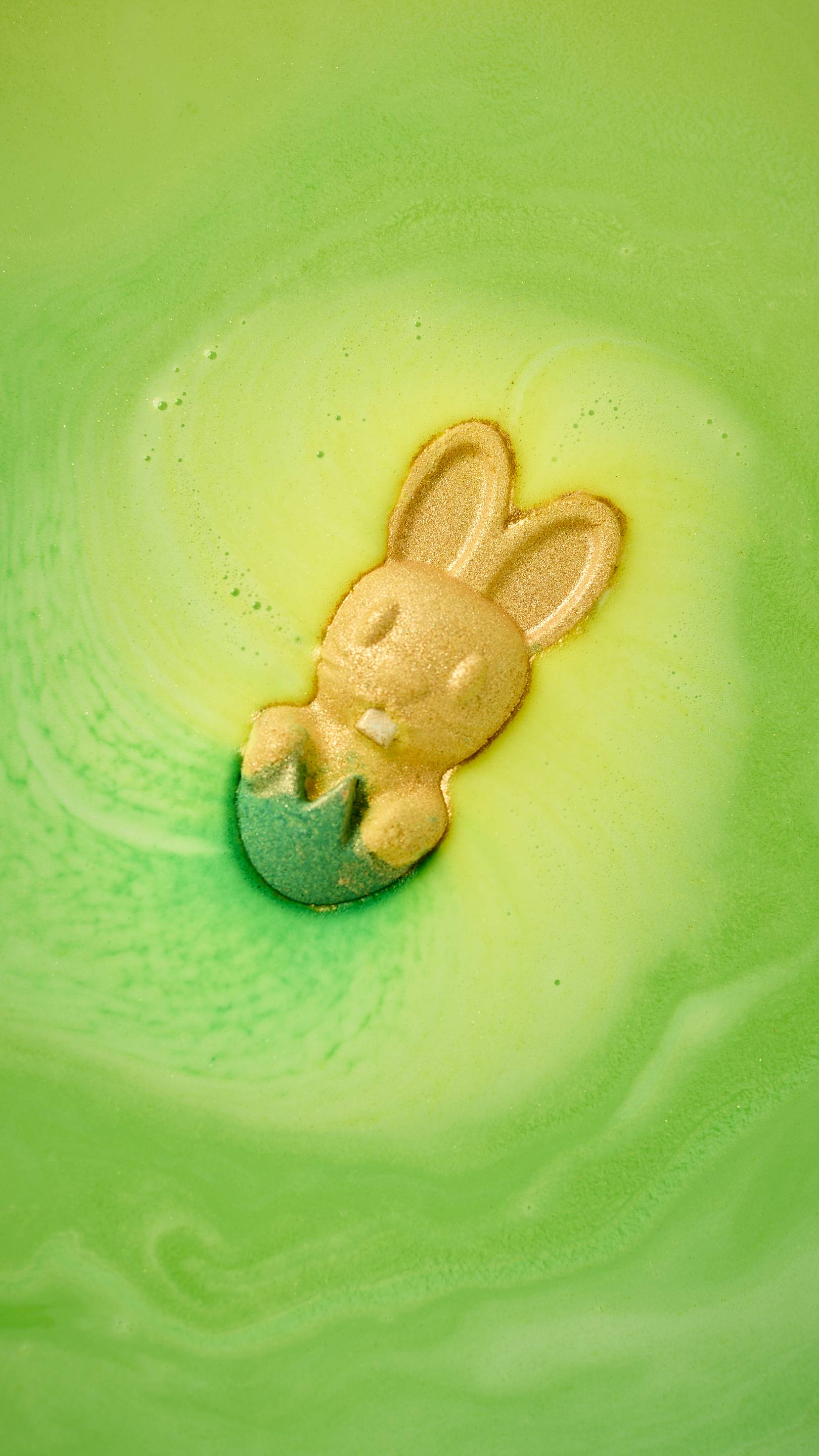 The Easter Bunny bath bomb, a golden rabbit popping out of a green grass-like base, floats in vibrant neon green bathwater. It releases swirling trails of bright yellow foam across the surface. All glitter used is plastic-free.