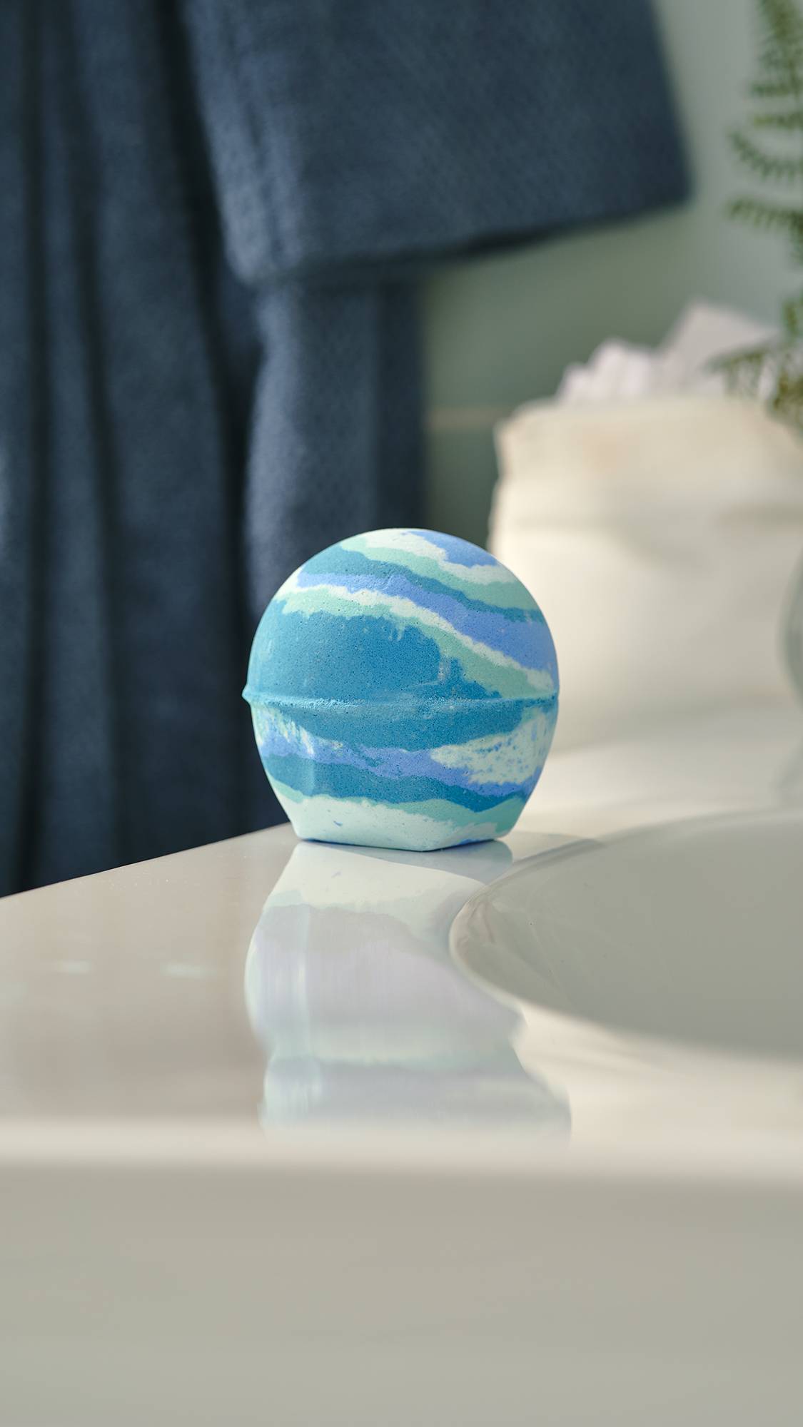 The image shows a spherical bath bomb with jagged horizontal stripes in various shades of blue and teal. It sits on the edge of a white ceramic bathroom sink, reflecting slightly on the surface with a dark blue towel blurred in the background.