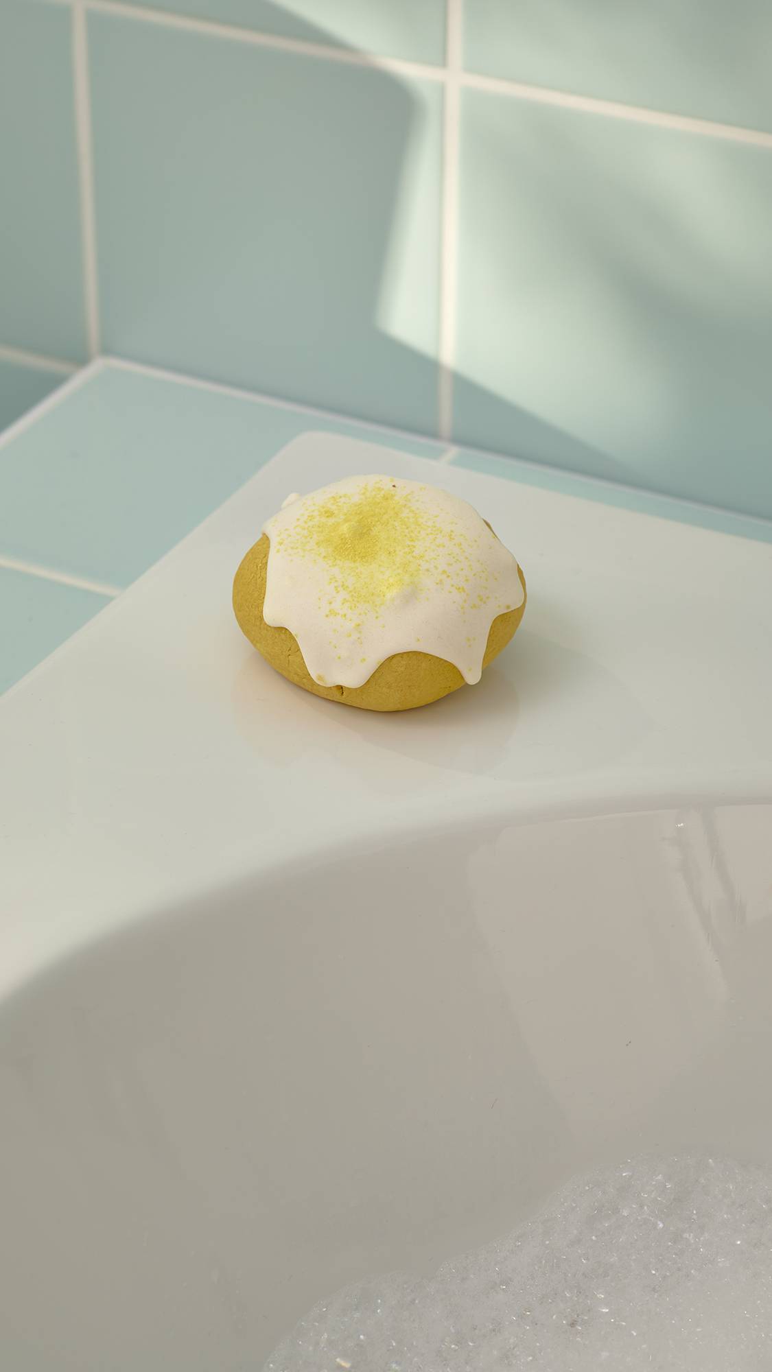 The image shows the "Easy Peasy Lemon Squeezy" bubble bar sitting on the corner of a white ceramic bathtub. The top is covered with a thick white drip and a dusting of yellow crystals, set against a background of pale mint green wall tiles.