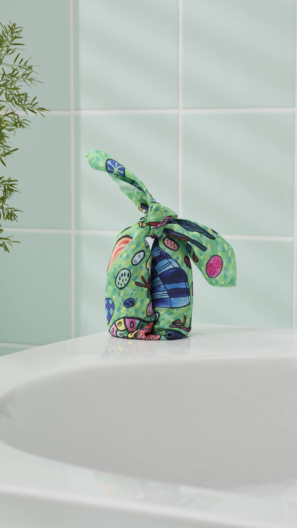 The image shows the Eggs For The Exotic Birds fabric knot wrap tied into a tall, bunny-ear knot around gifts. It stands on the edge of a white ceramic bathtub against a backdrop of pale mint-green wall tiles and a leafy green plant.