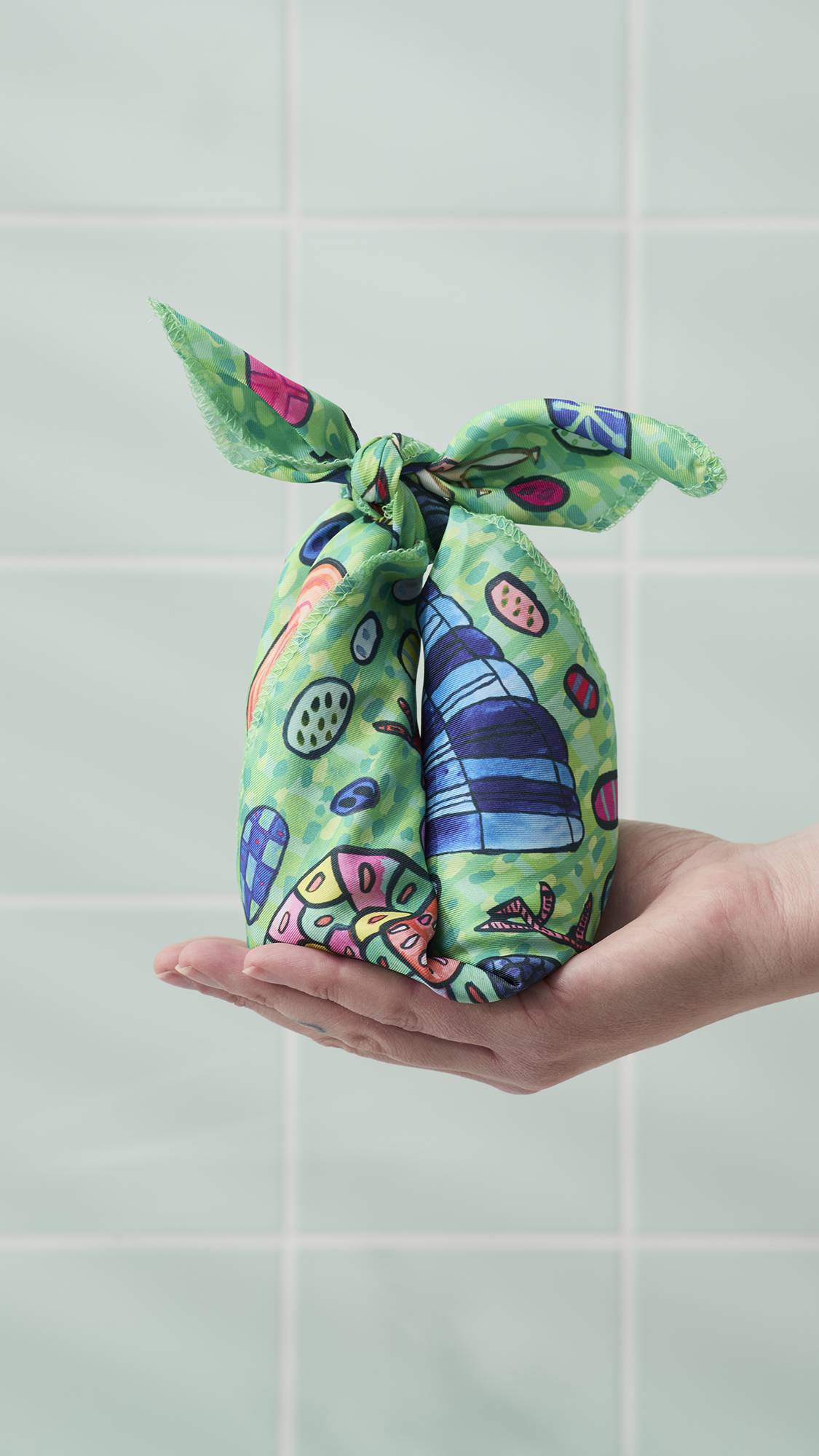 A hand holds an upright parcel in their palm, tied with the "Eggs For The Exotic Birds" knot wrap in a decorative "bunny ear" knot. The fabric wrap features a colourful hand-drawn pattern of birds and eggs, set against a background of bathroom tiles.