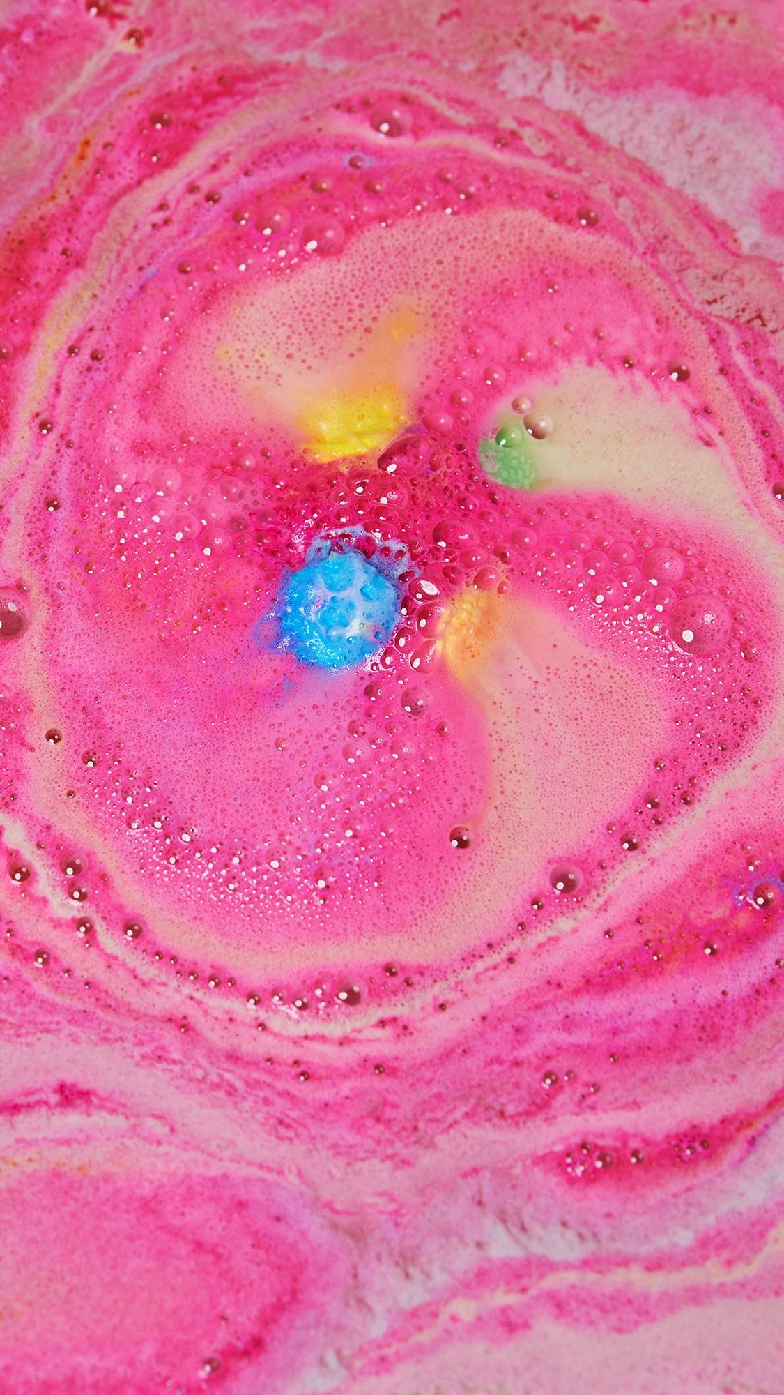 The bright pink egg part of the bath bomb sits spinning in the water giving off hot pink foam with flashes of yellow, green and blue. 