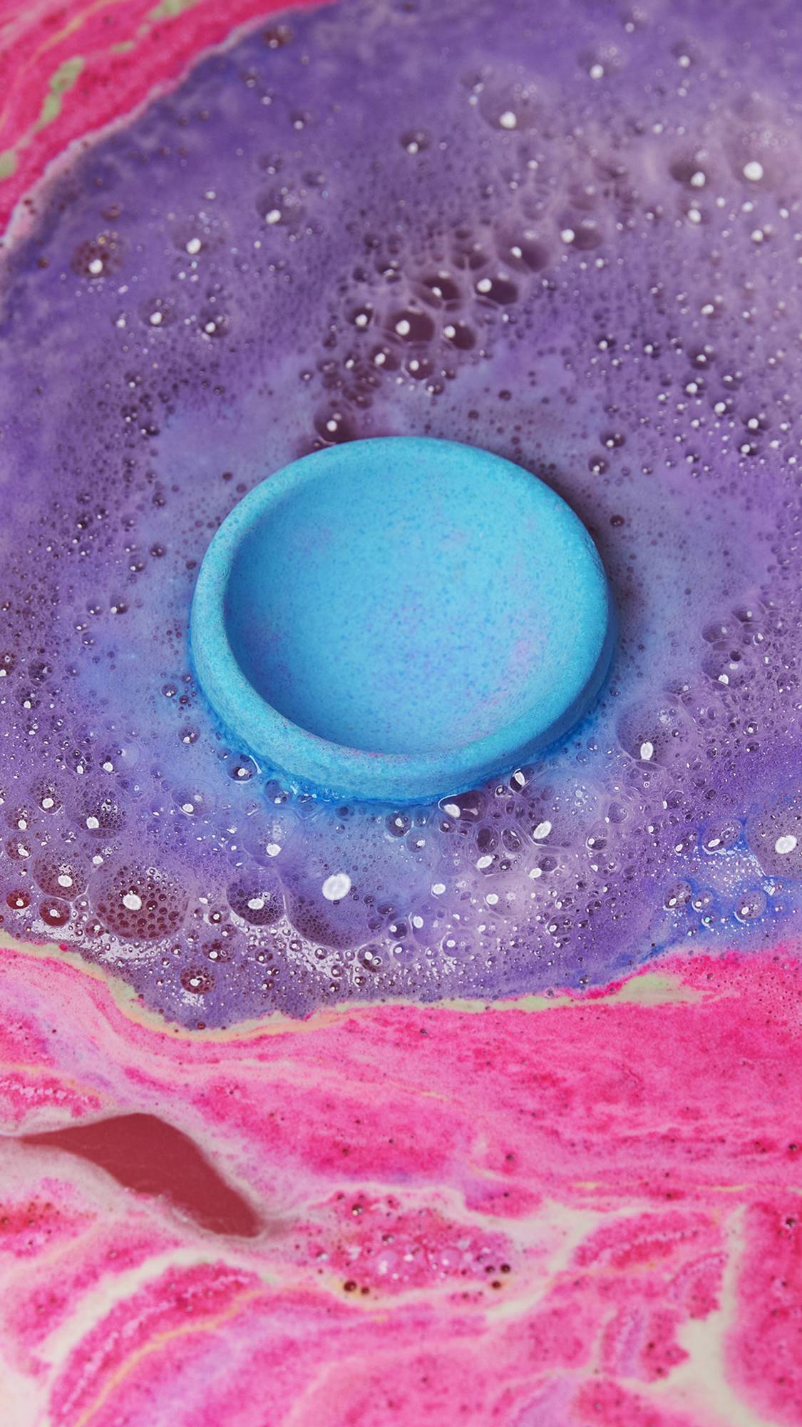 The egg cup part of the bath bomb sits in the water giving off a deep purple foam while surrounded by pink water from the egg. 