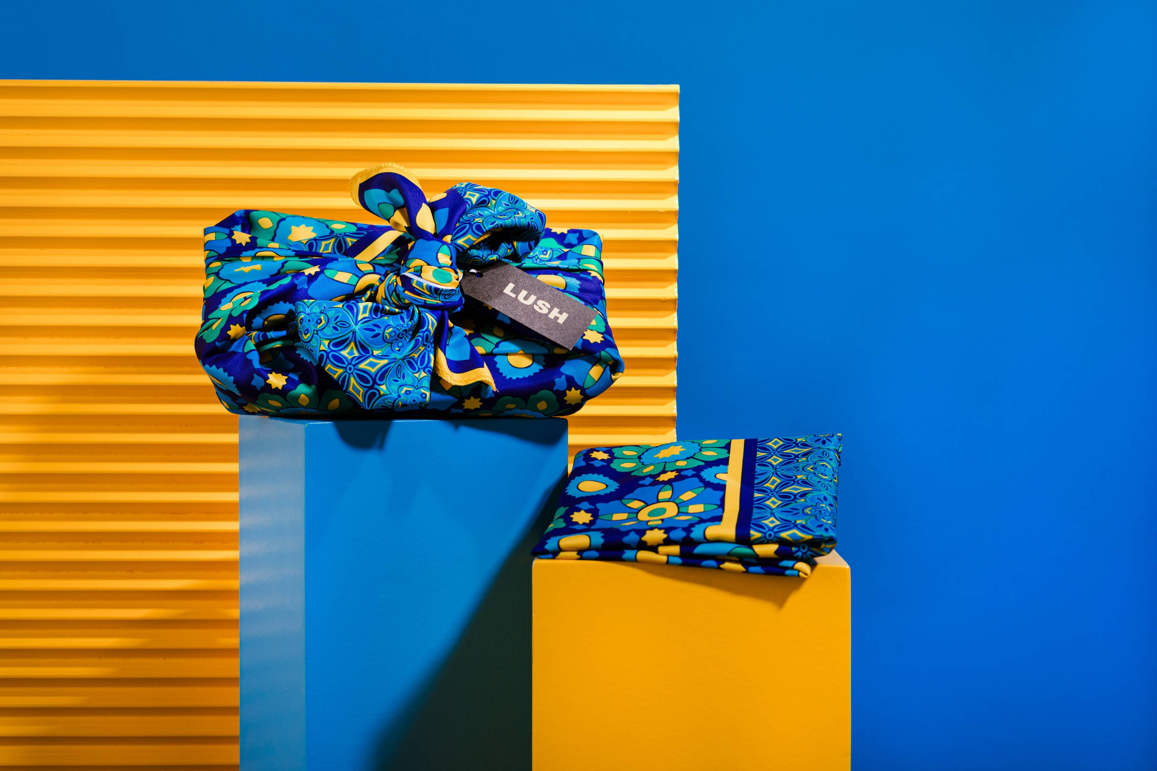 The Knot Wrap is decoratively wrapped with a Lush gift tag, alongside one folded, in front of a blue and yellow background.