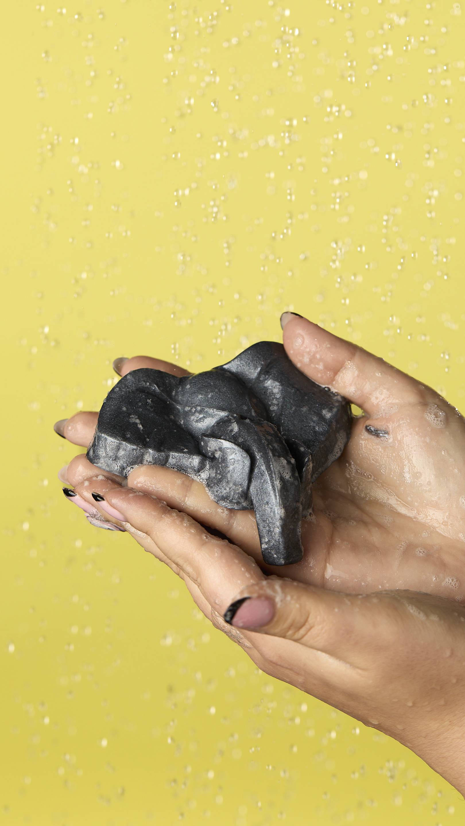 The image shows a pair of light-toned hands reaching in from the right. They are cupped and gently holding the metallic silver Elephant Jelly in their hands. The background is a solid yellow with blurred water droplets falling. 