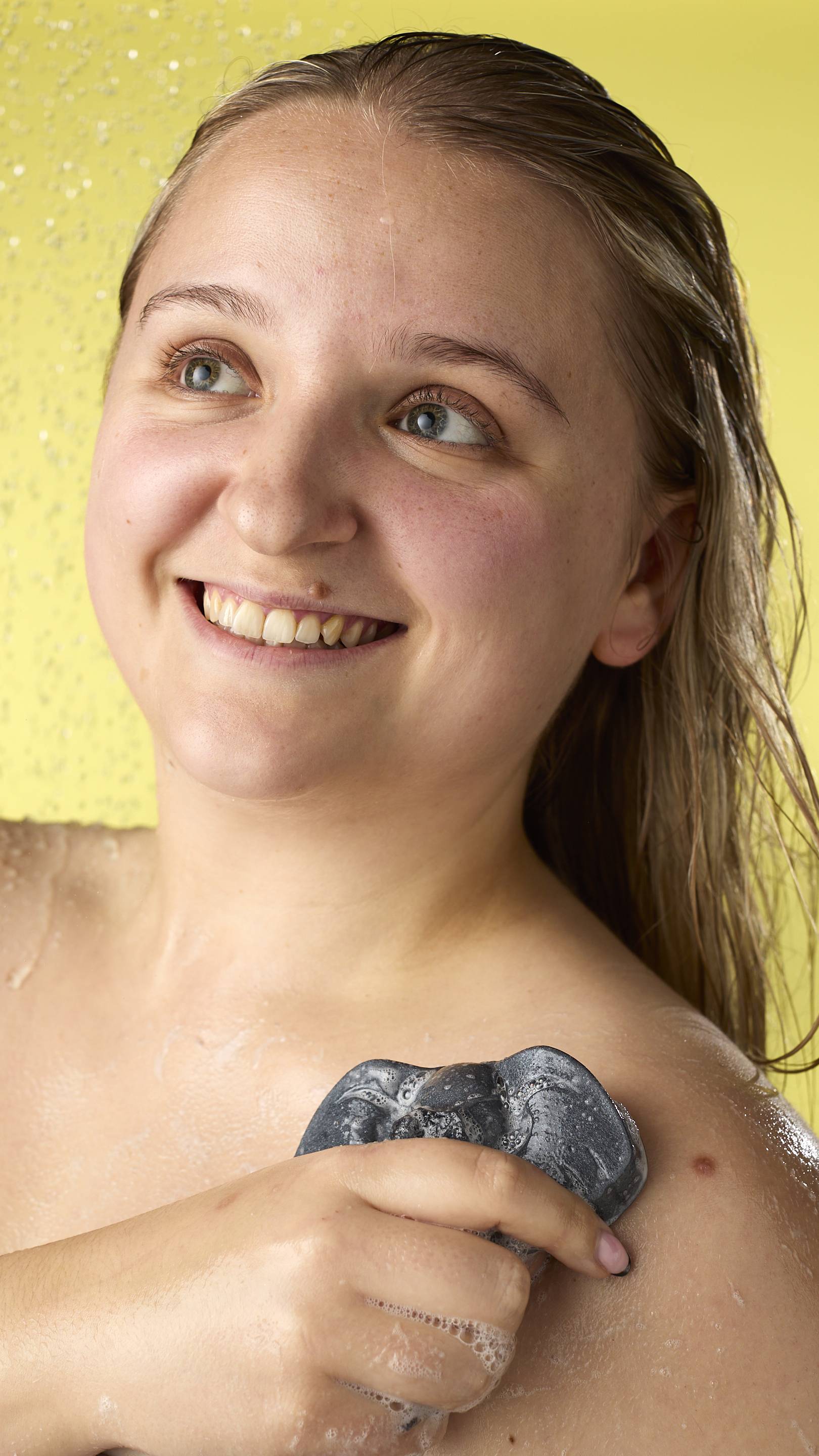 The image shows a blonde, smiling model in the shower, from the shoulders up. They are using a metallic silver elephant jelly to wash their shoulder. Water droplets and suds are visible on their skin, and the background is a solid bright yellow.