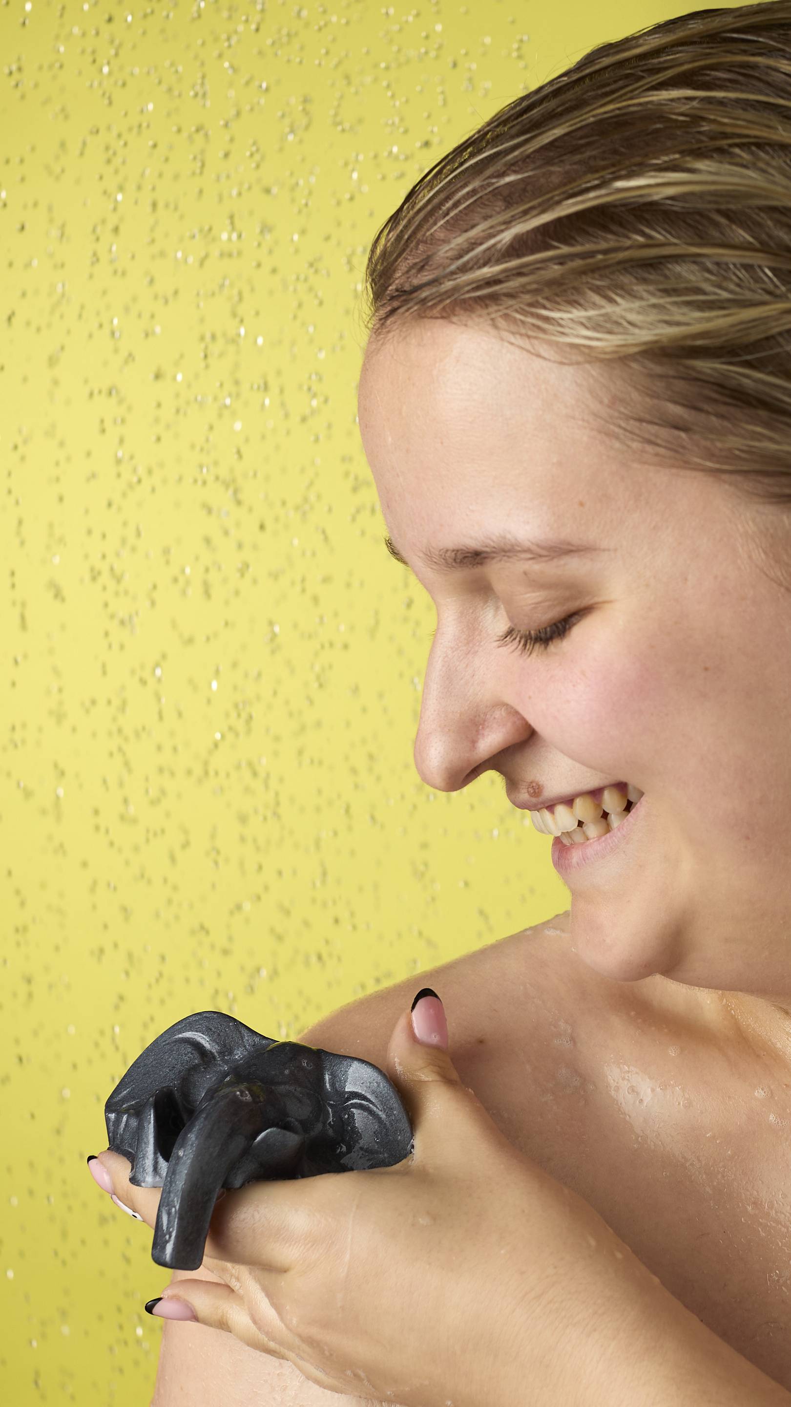The image shows a blonde, smiling model in the shower, from the shoulders up. They are using a metallic silver elephant jelly to wash their shoulder. Water droplets and suds are visible on their skin, and the background is a solid bright yellow.
