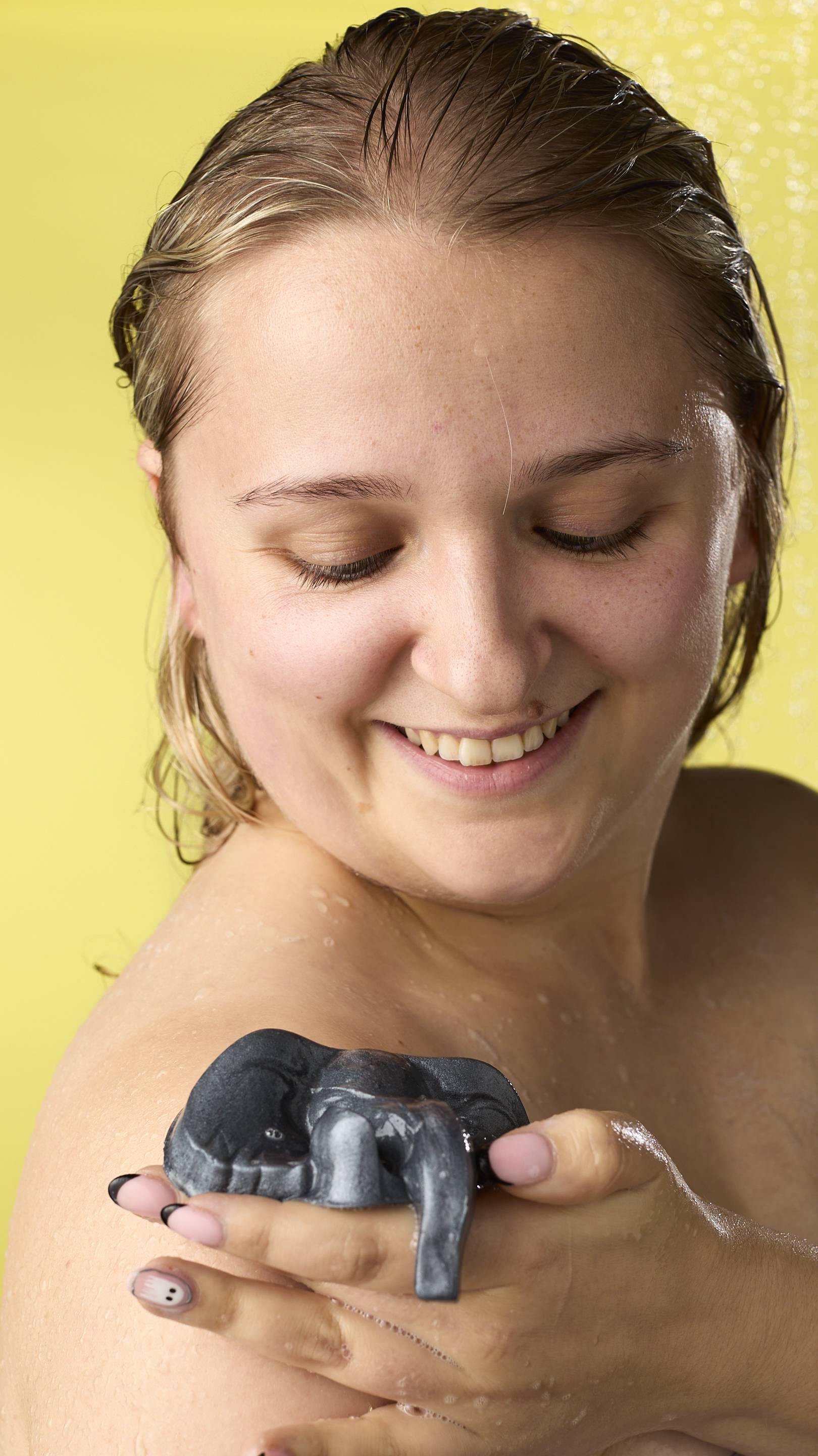 The image shows a blonde, smiling model in the shower, from the shoulders up. They are using a metallic silver elephant jelly to wash their shoulder. Water droplets and suds are visible on their skin, and the background is a solid bright yellow.
