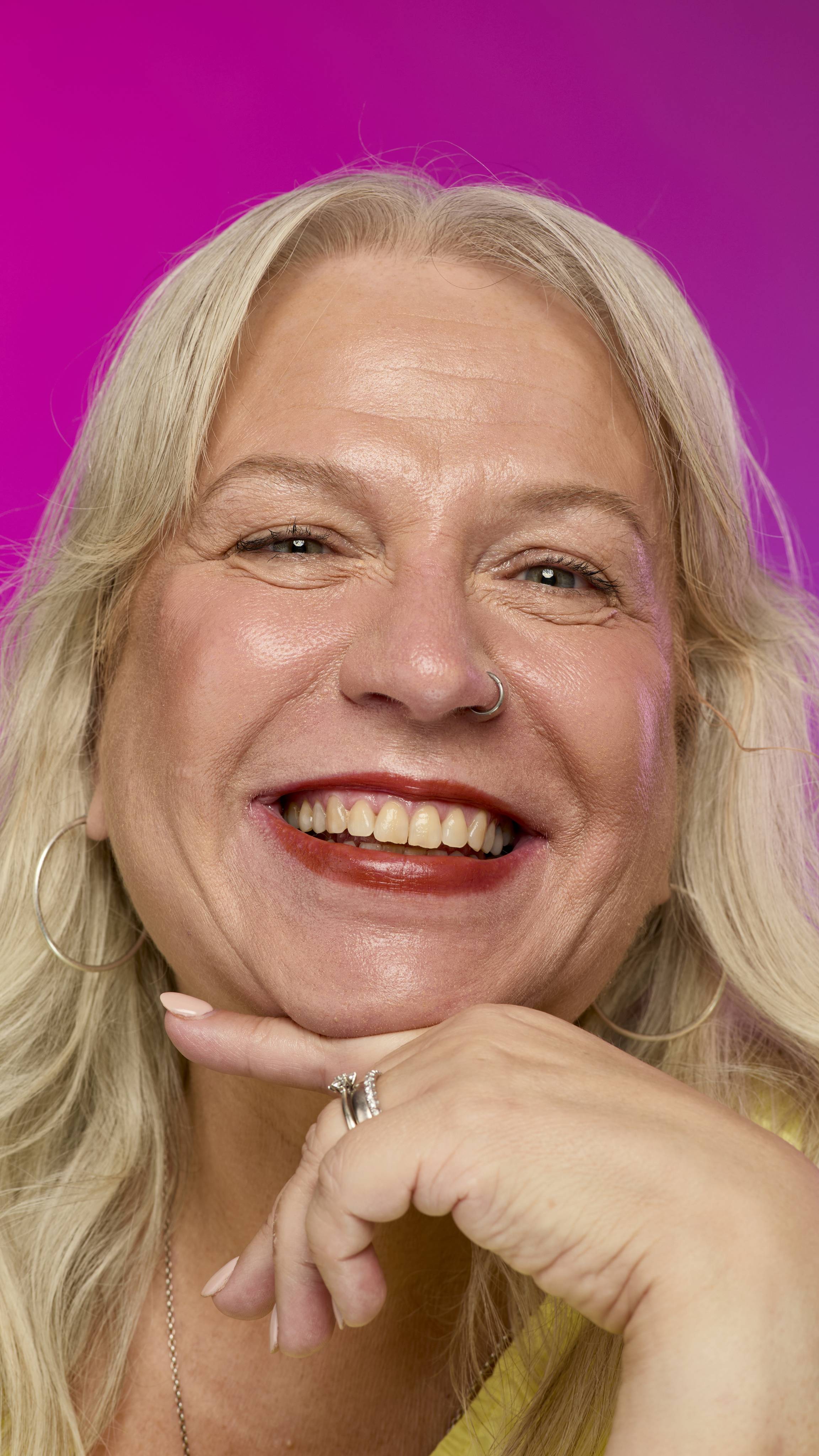 A smiling model with fair skin and light hair is shown from the neck up, with their chin resting on their hand, on a vibrant pink background. They smile, showing off the rich, mahogany Ember lipstick with a subtle shimmer. 