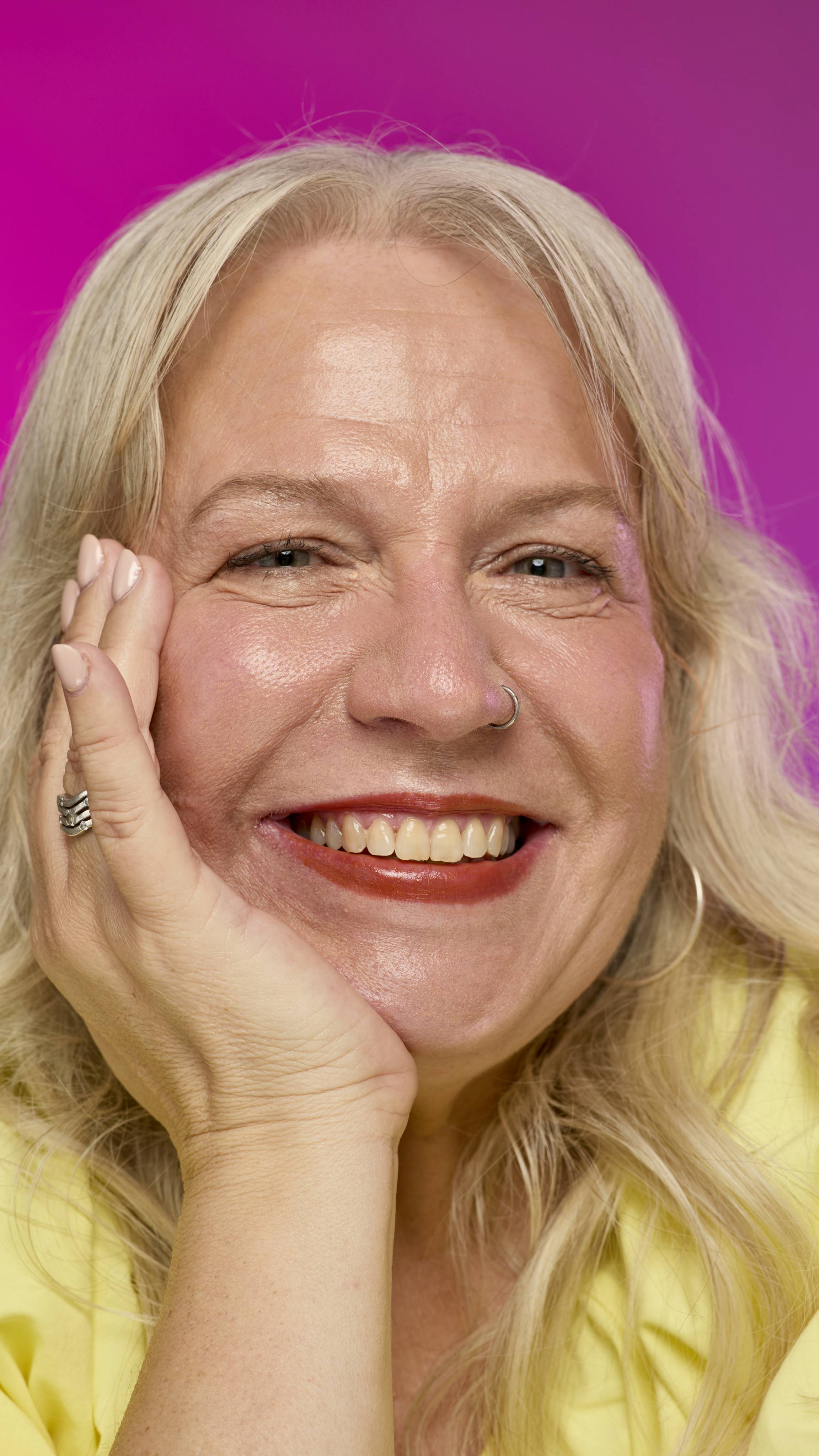 A smiling model with fair skin and light hair is shown from the neck up, with their chin resting on their hand, on a vibrant pink background. They smile, showing off the rich, mahogany Ember lipstick with a subtle shimmer. 