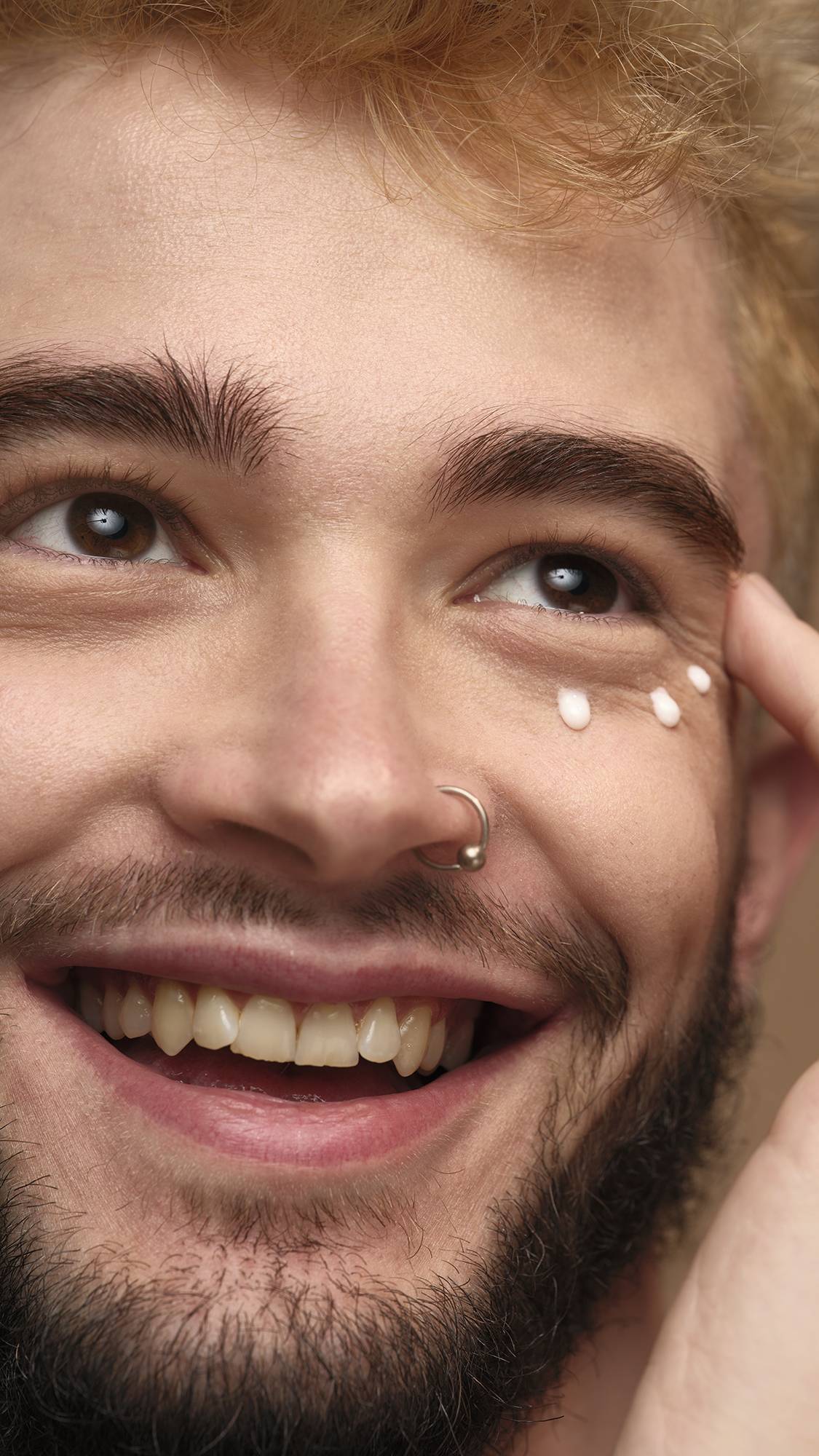 A close-up of the model's face. They are smiling with three drops of Enchanted eye cream neatly spotted under their eyes as they are about to smooth it into their skin.