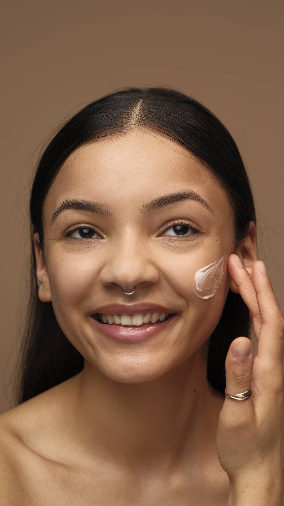 An image of the model's smiling face as they start to apply the Enzymion self-preserving moisturiser to one cheek. They are on a warm, earthy-brown background. 