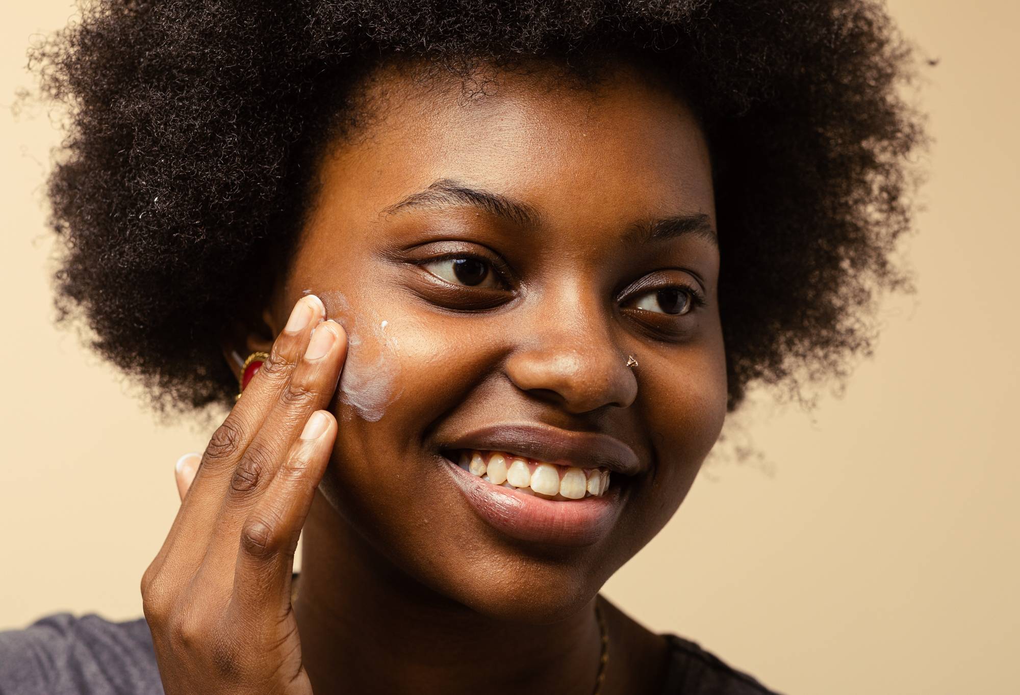 A person with coily, afro hair smiles as they rub a small patch of cream coloured Enzymion moisturiser into their cheek.