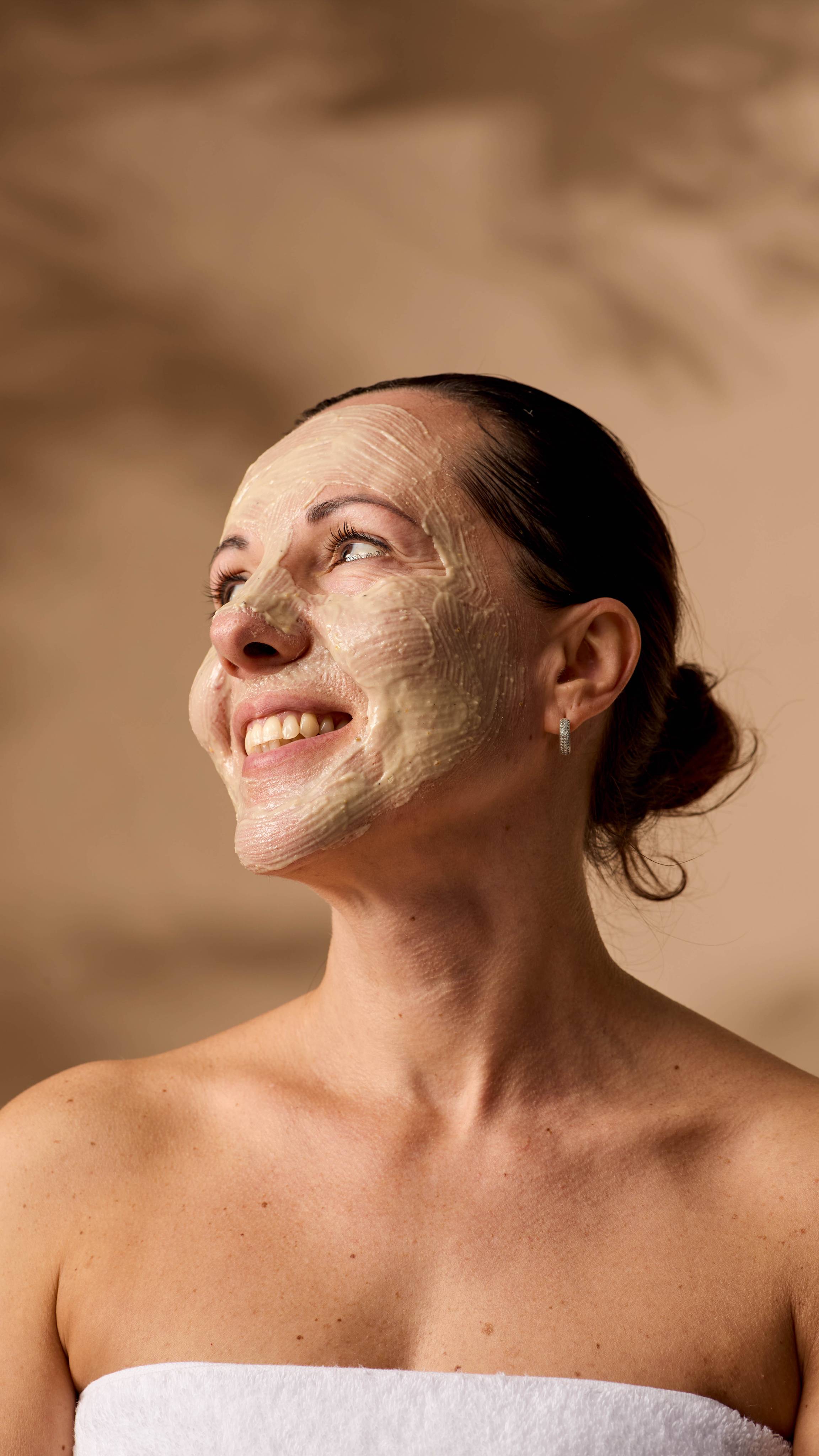 The model is wrapped in a fluffy, white towel, smiling into the distance with their face evenly coated in the Enzynamite face mask.