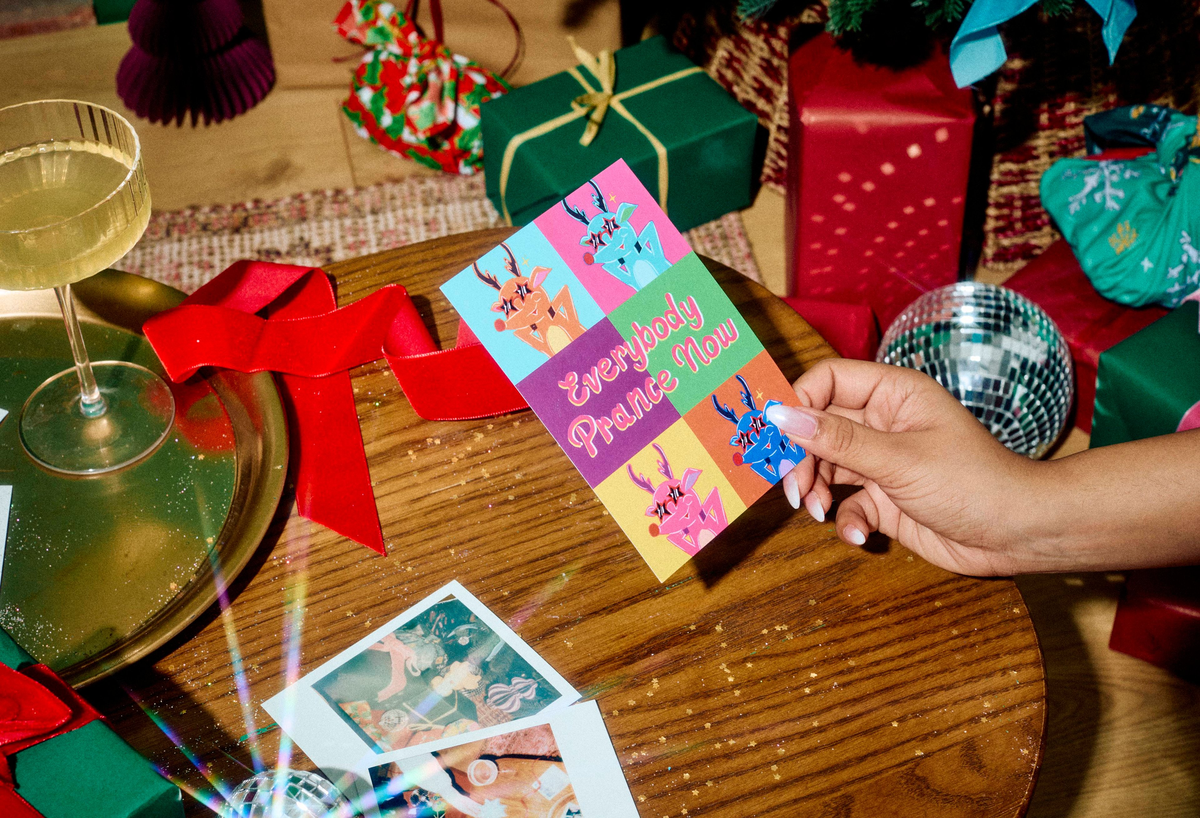 A hand holds a pop-art style Lush postcard reading "Everybody Prance Now" over a wooden table set with drinks, disco balls, red ribbon, and wrapped festive gifts.