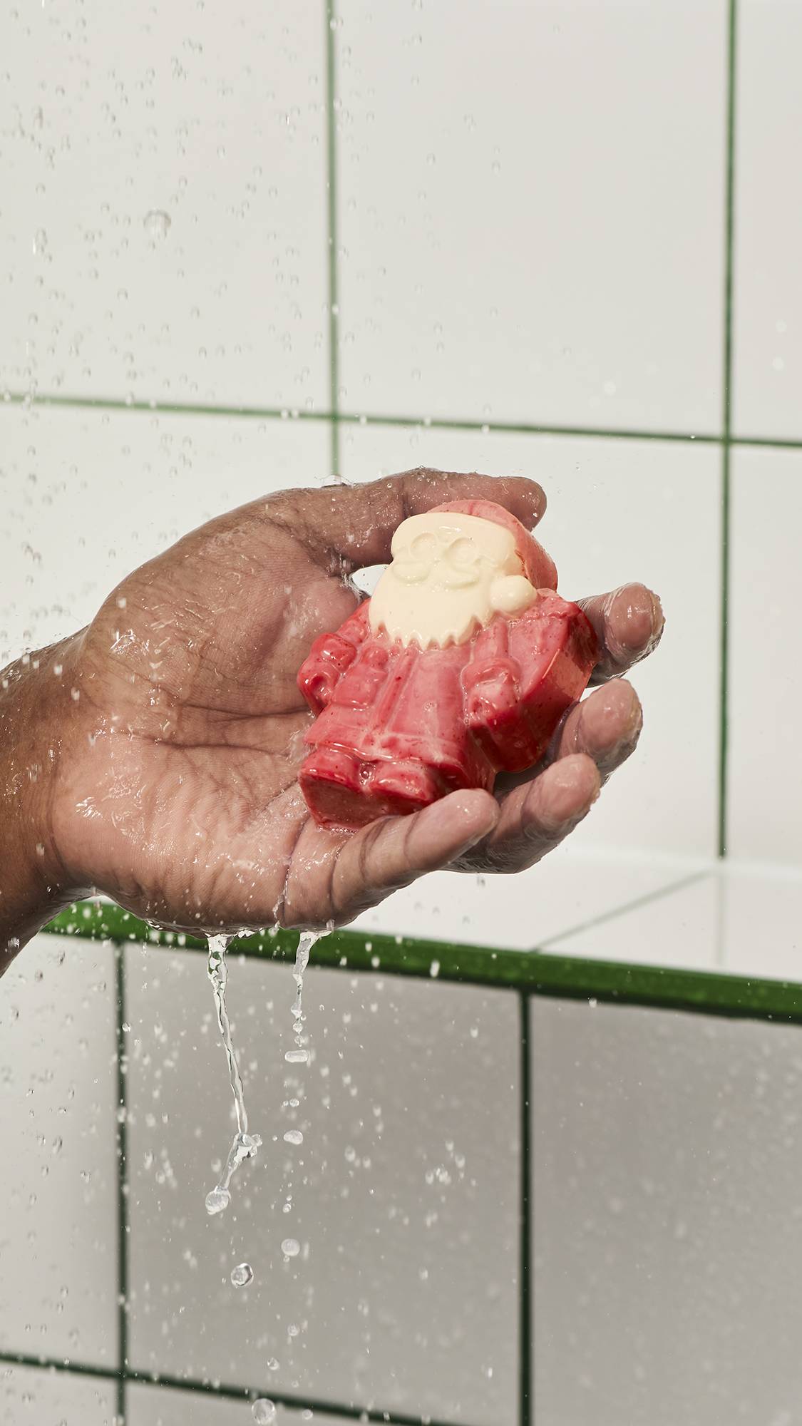 A hand holds a small moulded Santa Claus-shaped soap bar with a red suit and cream beard under streaming shower water, set against a white tiled wall background.