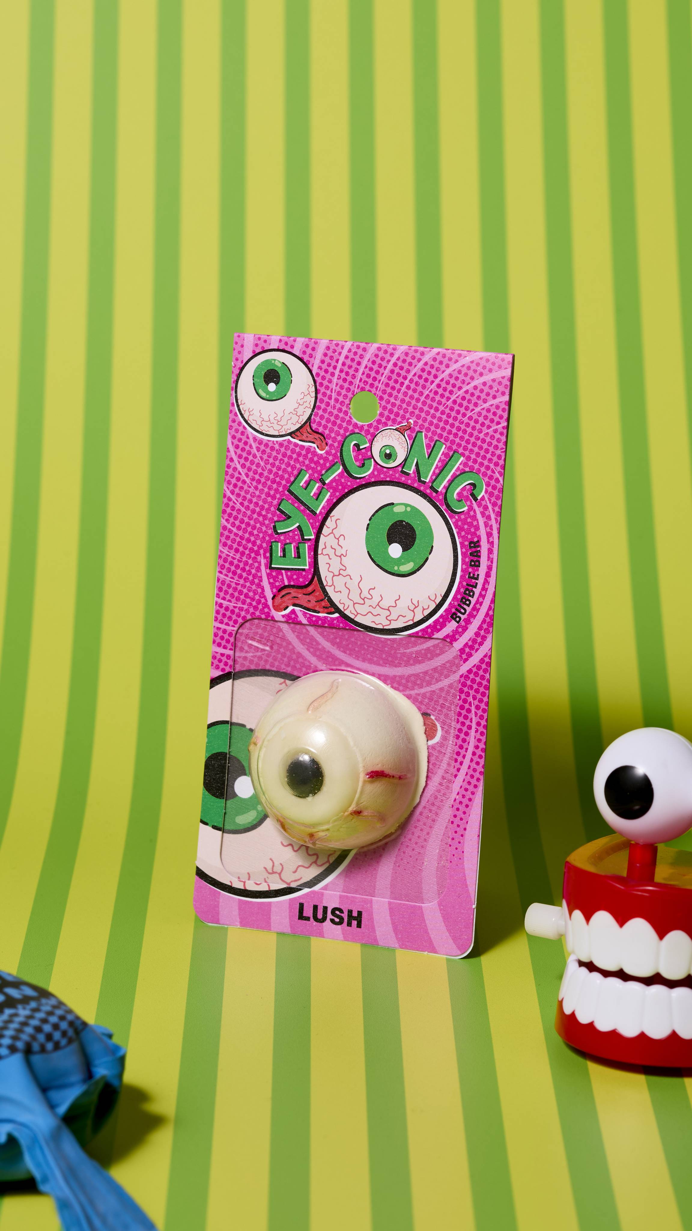 The image shows the eyeball-shaped, Eye-Conic bubble bar product on a yellow and green striped background. There is a whoopie cushion and wind-up chattering teeth that are just visible.