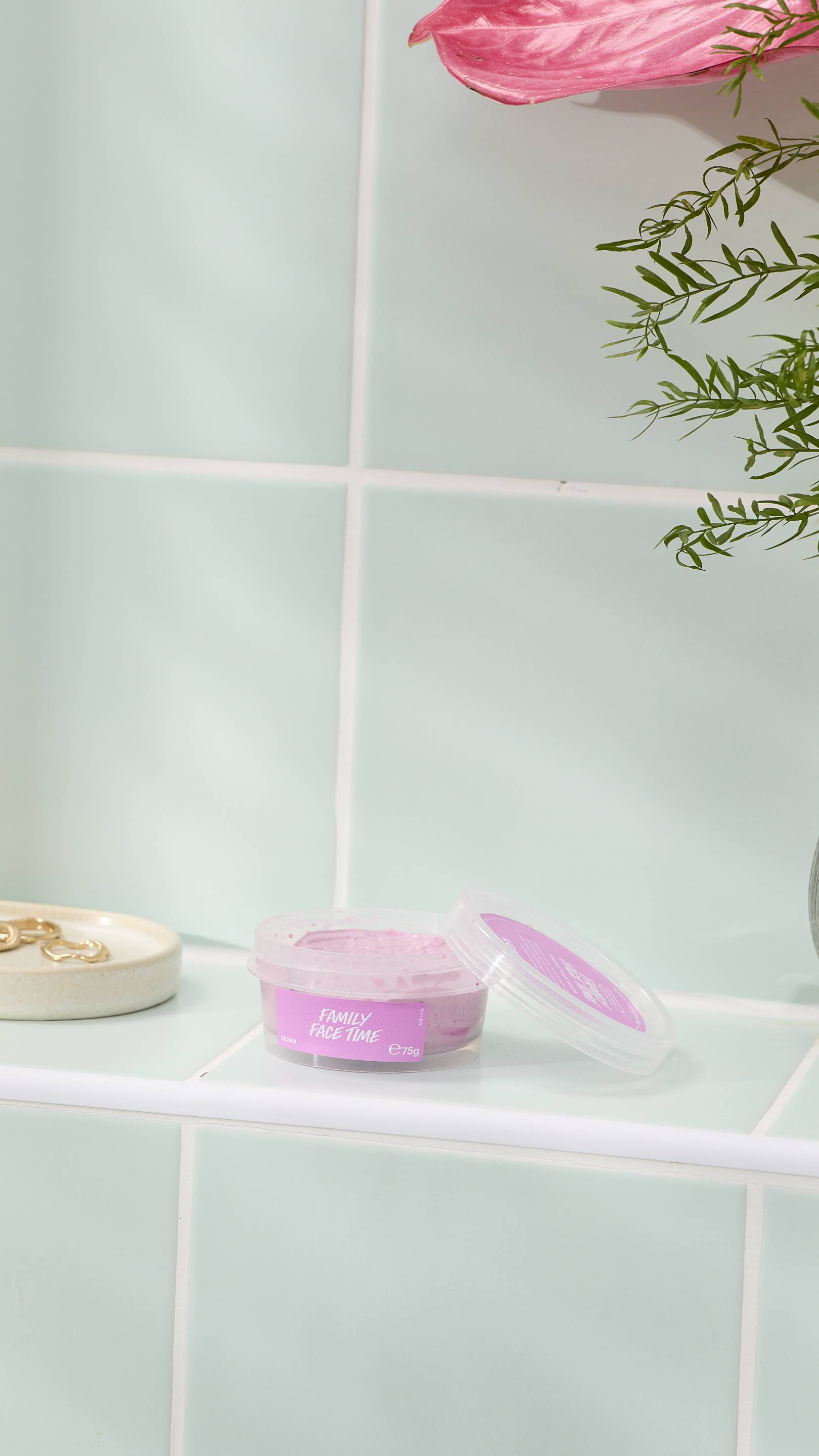 An open, clear plastic pot of 'FAMILY FACE TIME' fresh face mask sits on a pale green tiled ledge. The pot contains a thick, grainy pink face mask, with some residue on the lid. A small dish with gold rings and a pink flower are nearby.