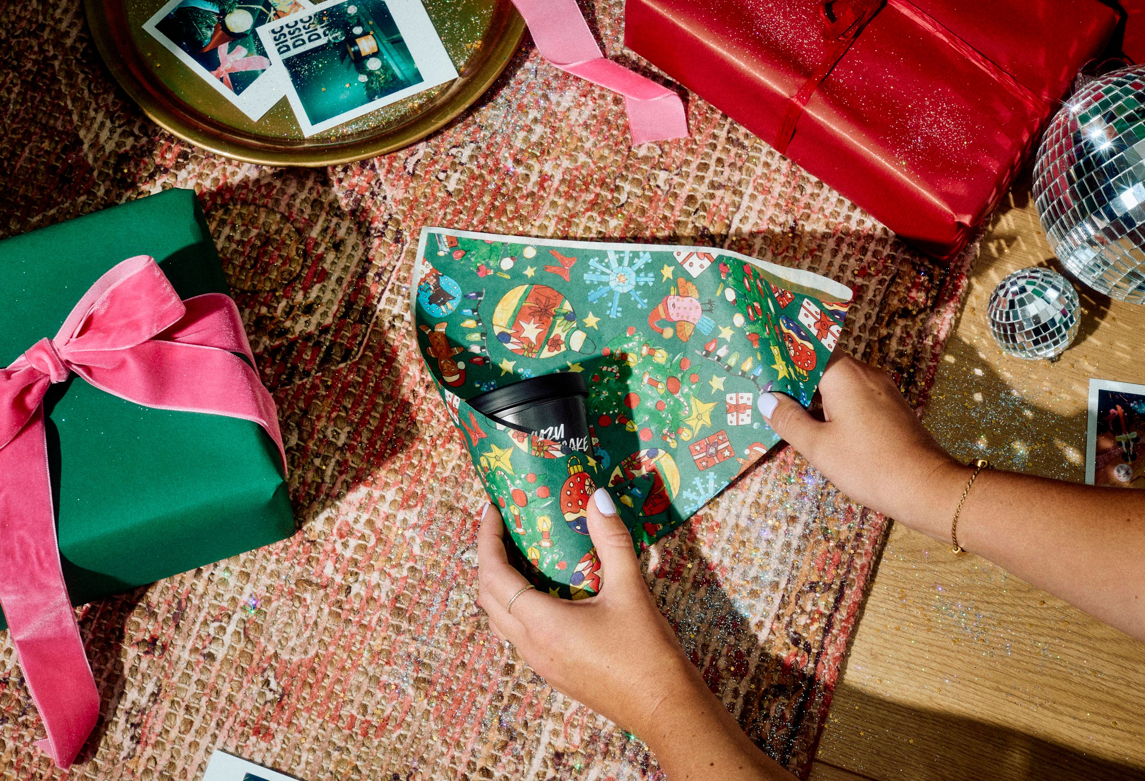 Hands wrap a black pot using a green festive-patterned sheet of Lokta wrapping paper, viewed from above on a patterned rug surrounded by wrapped presents, disco balls, and instant photos.