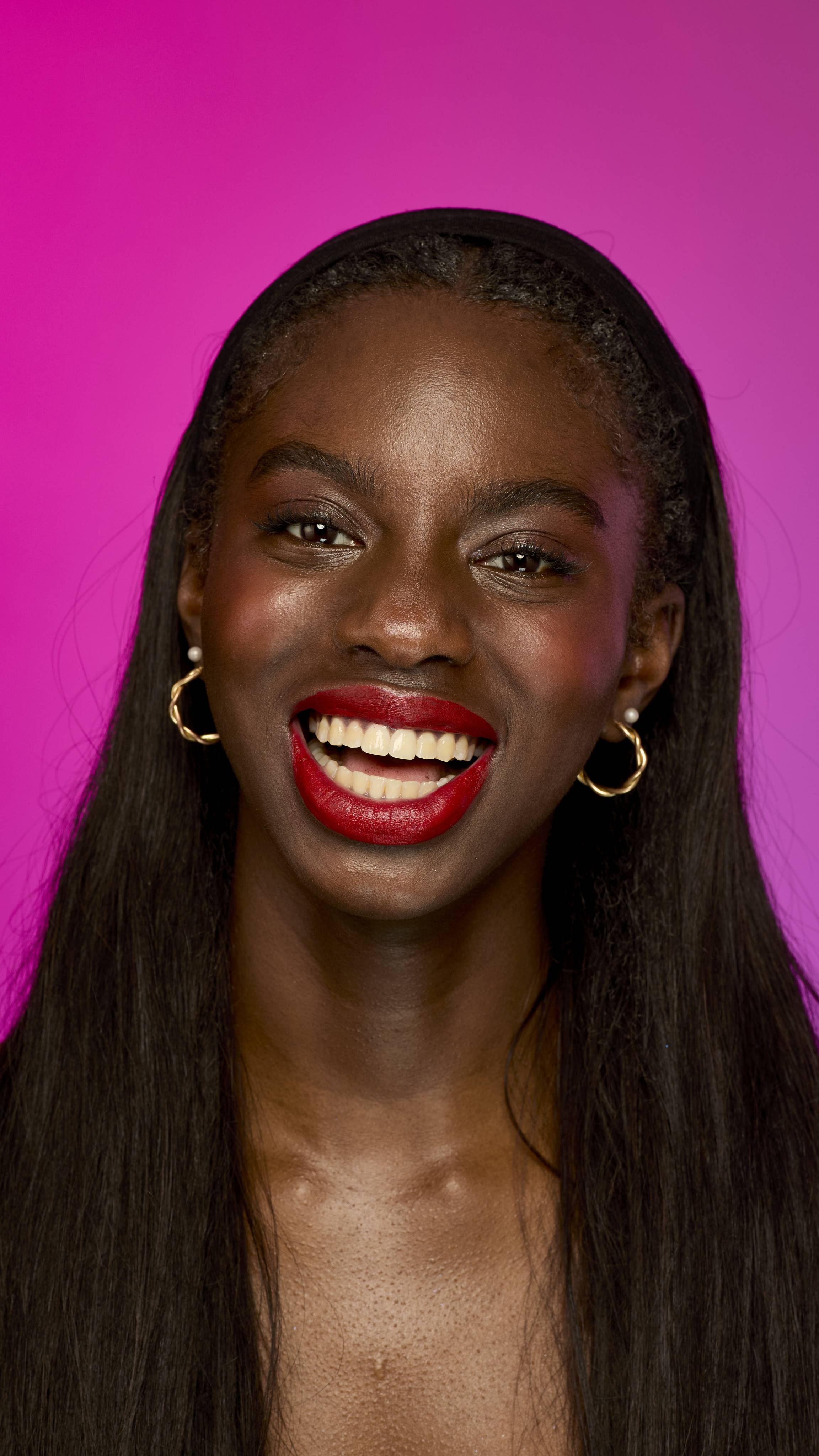 The image shows a model with long, dark hair on a vibrant magenta background. They are smiling widely, showing the scarlet-red Firecracker lipstick.