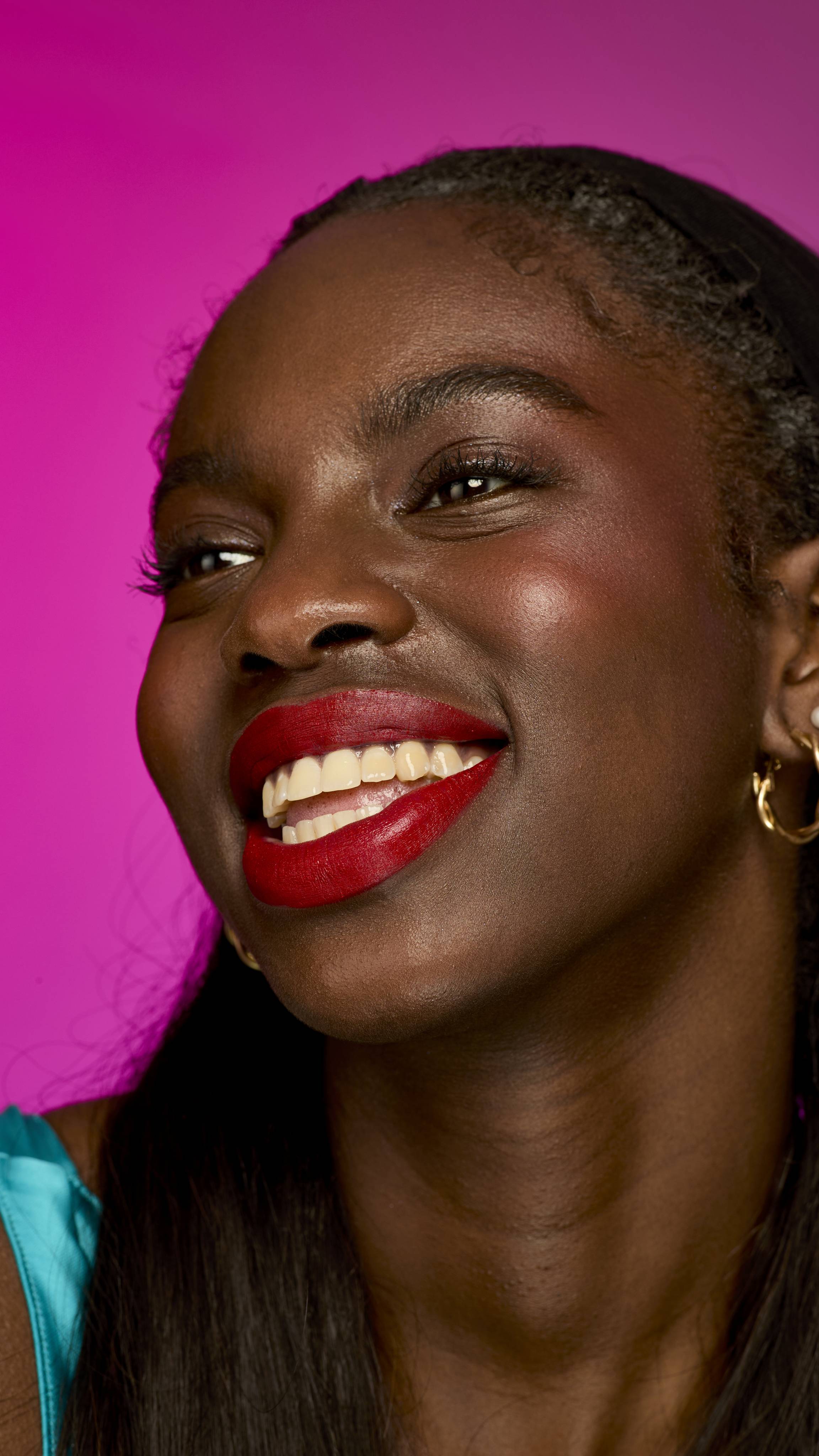 The image shows a model with long, dark hair on a vibrant magenta background. They are smiling widely, showing the scarlet-red Firecracker lipstick.