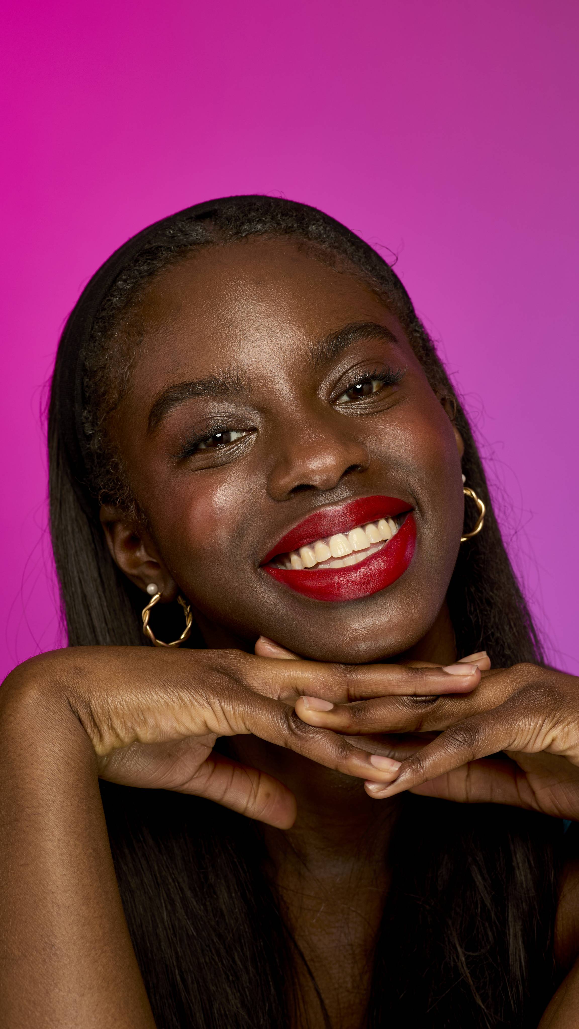 The image shows a model with long, dark hair on a vibrant magenta background, holding their chin up with interlocked hands. They are smiling widely, showing the scarlet-red Firecracker lipstick.