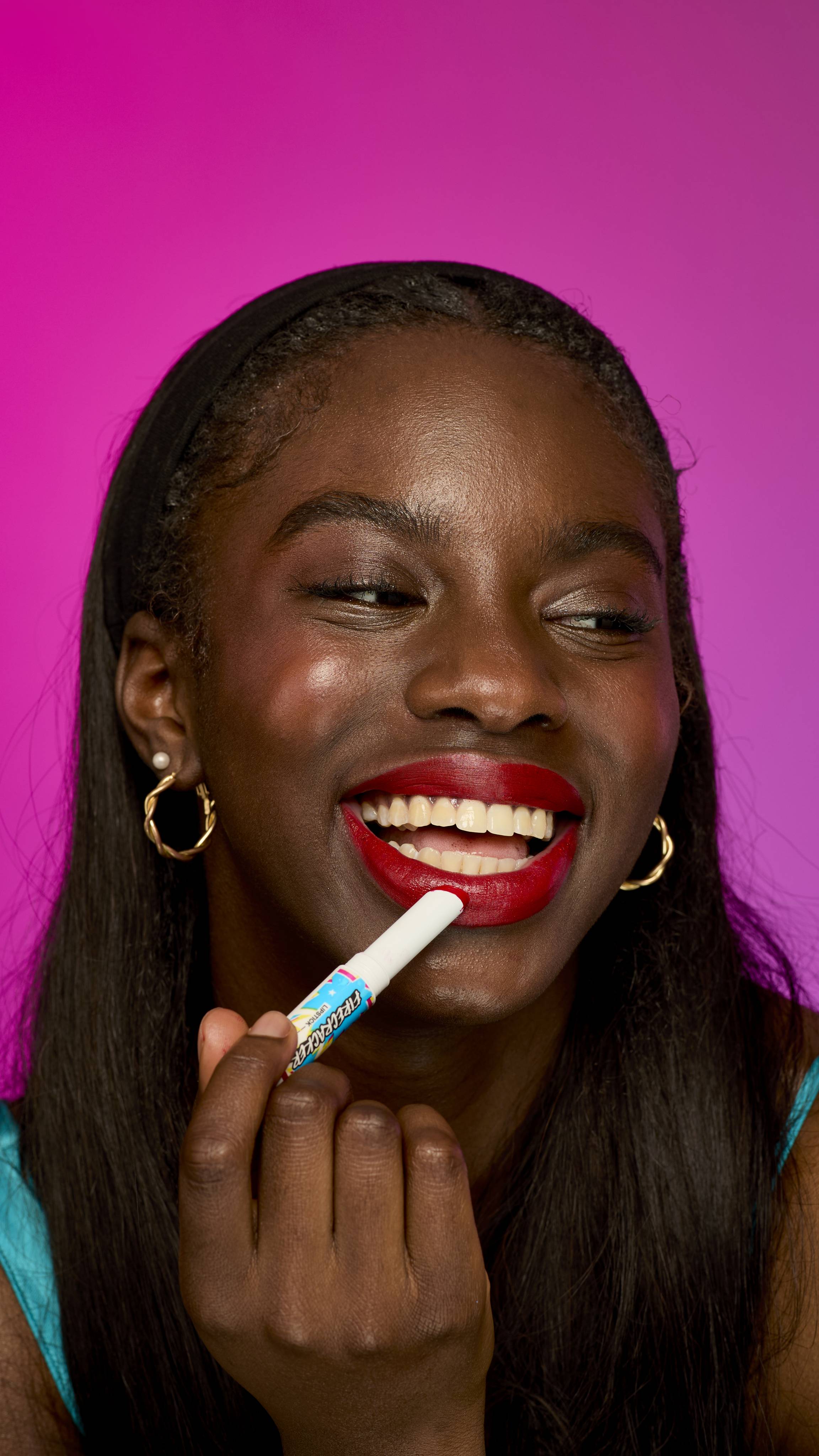 A model with long, dark hair is shown from the chest up on a vibrant, solid magenta background. They are gently applying the velvety, scarlet-red twist-up Firecracker lipstick.