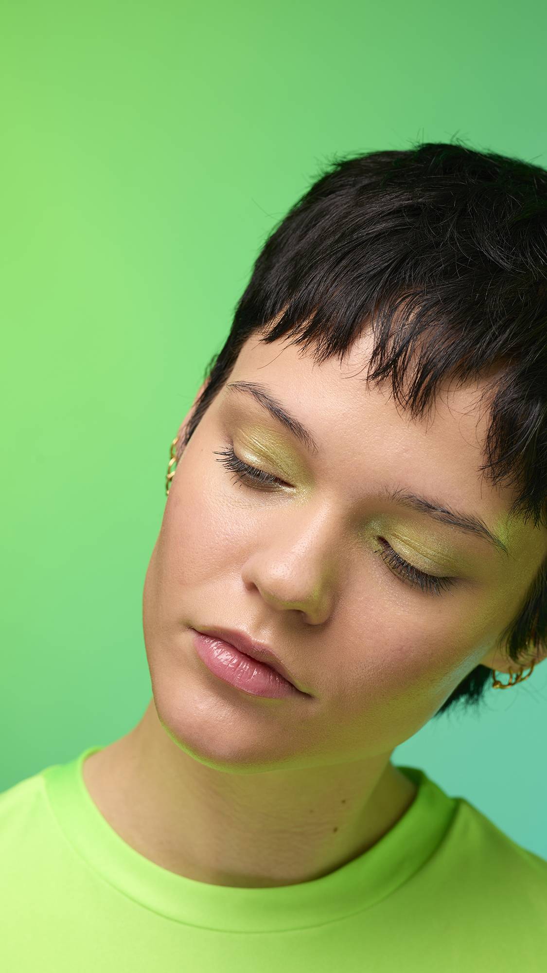  The image shows a close-up of the model's face on a pale blue-to-green gradient background. The model's make-up is done with the focal point being a subtle, lime-green, shimmering eyeshadow made by the Firefly Glitter Crumble. 