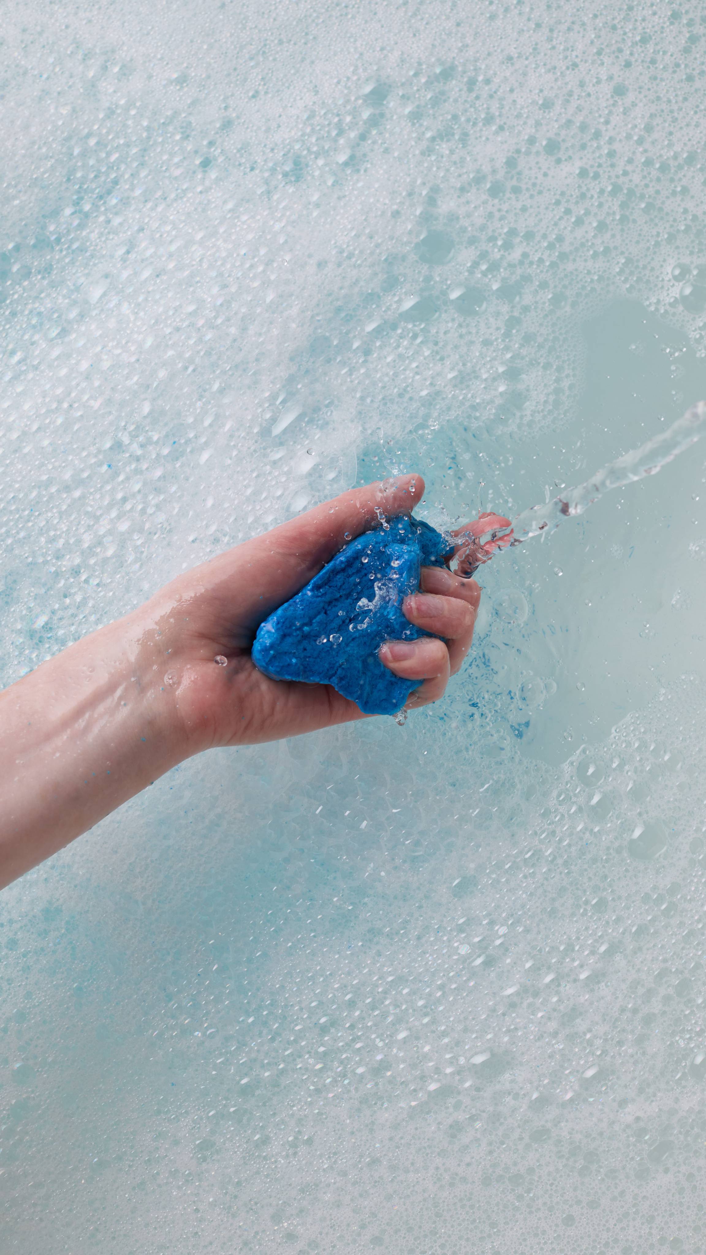 The image shows the model's hand reaching in from the left as it crumbles the bright blue, First Swim bubble bar under running tap water in a white porcelain bath. The cool-blue-coloured water has a thick layer of bubbles forming.