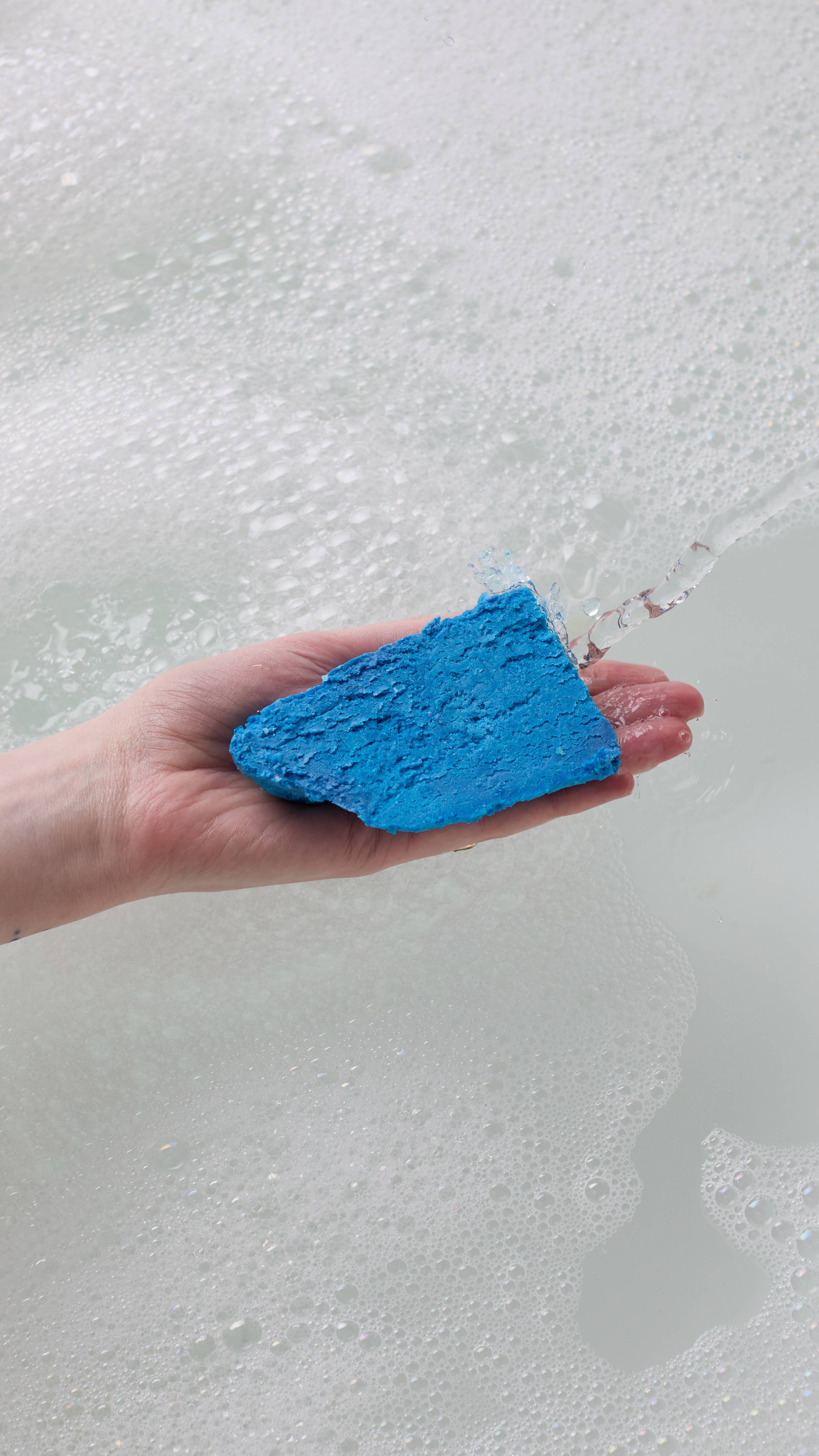 The image shows the model's hand reaching in from the left as it holds the bright blue, First Swim bubble bar in front of running tap water in a white porcelain bath. The water has a thick layer of bubbles forming.