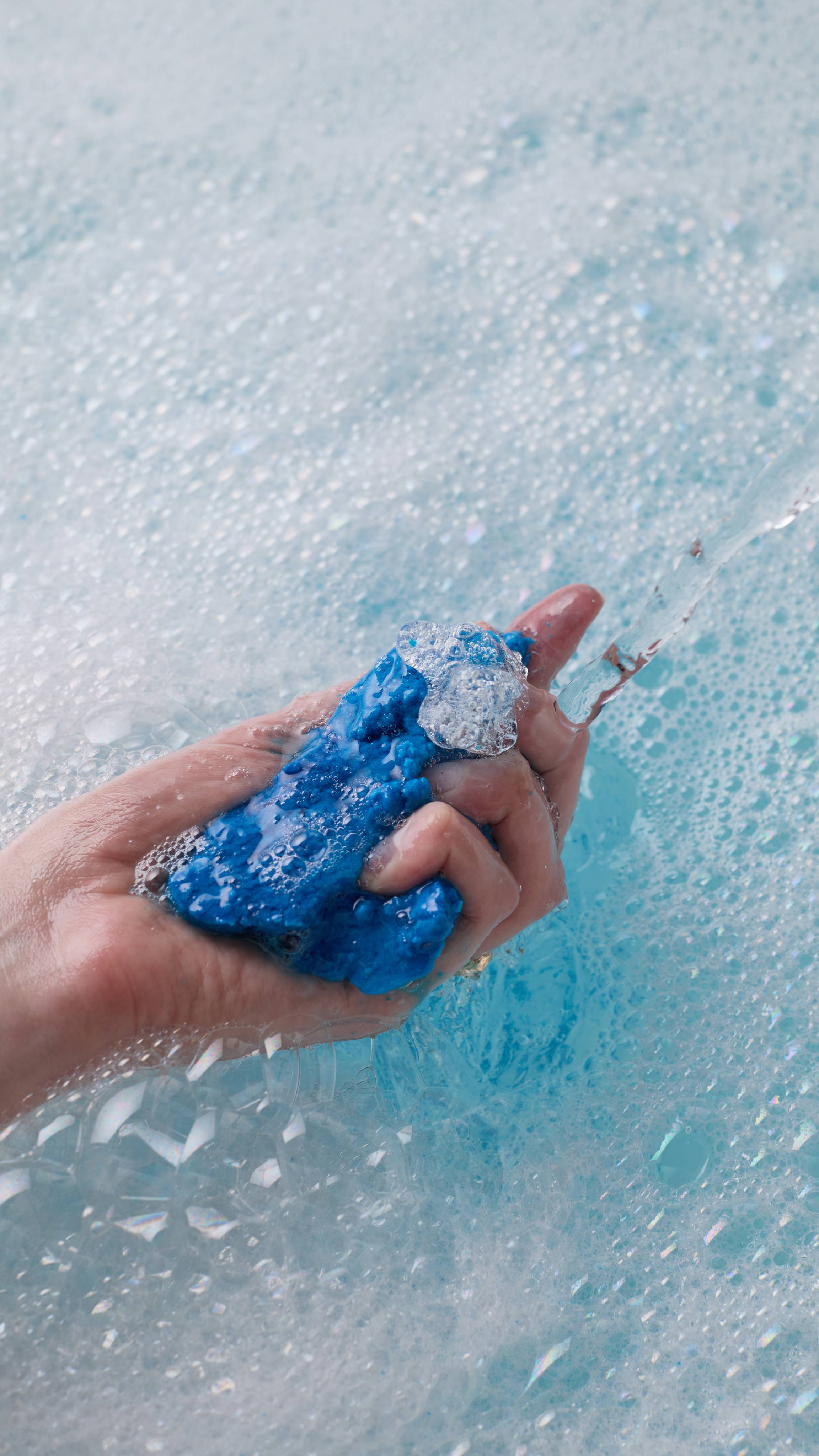 The image shows the model's hand reaching in from the left as it crumbles the bright blue, First Swim bubble bar under running tap water in a white porcelain bath. The cool-blue-coloured water has a thick layer of bubbles forming.
