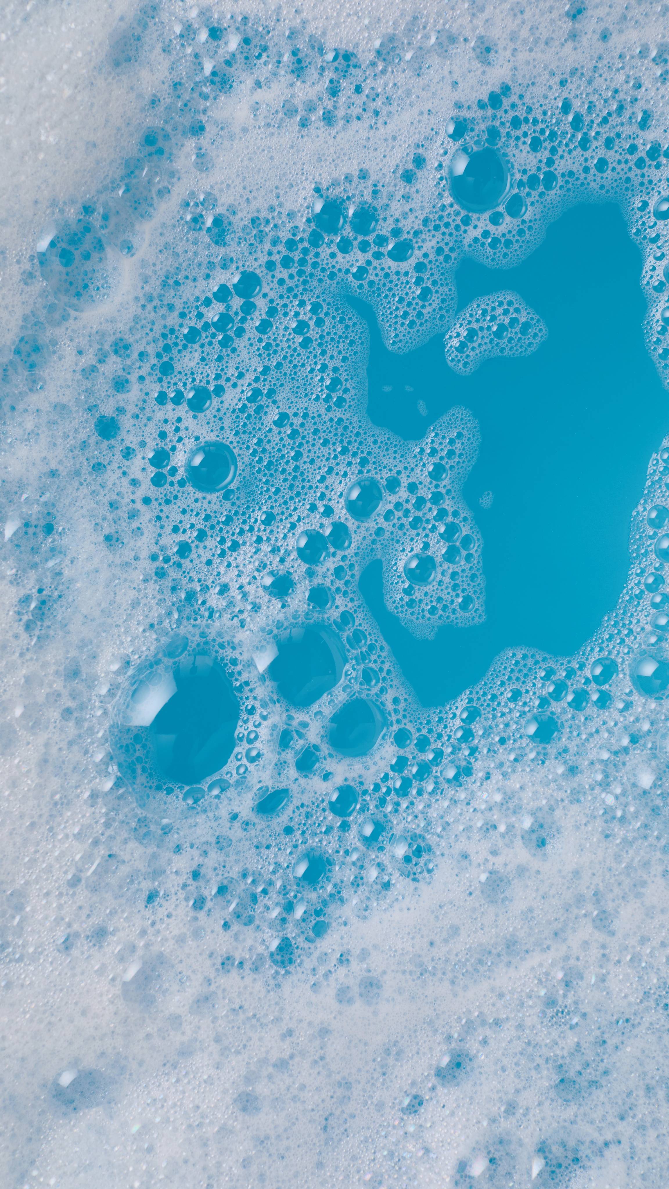 A close-up, aerial image of the bath water after using the First Swim bubble bar. There is a blanket of thick, fat bubbles with bright, icy, cool-blue bath water visible through a large gap.